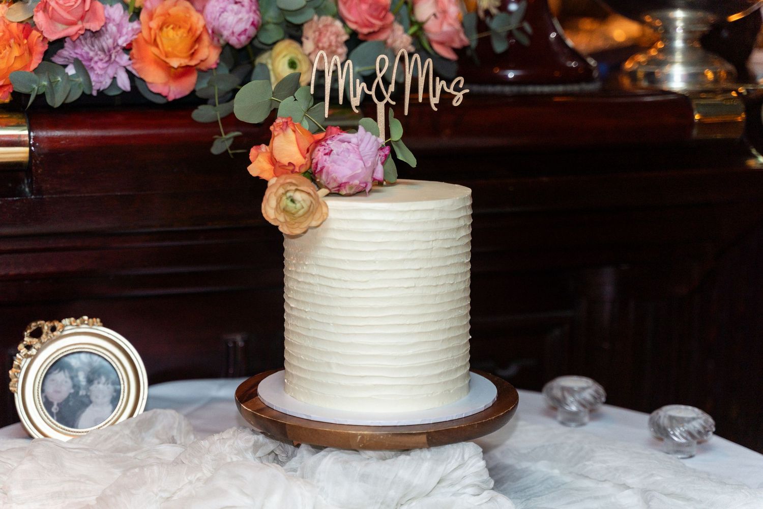 White ribbed single-tier wedding cake with Mr & Mrs topper and colorful floral arrangement on wooden cake stand.