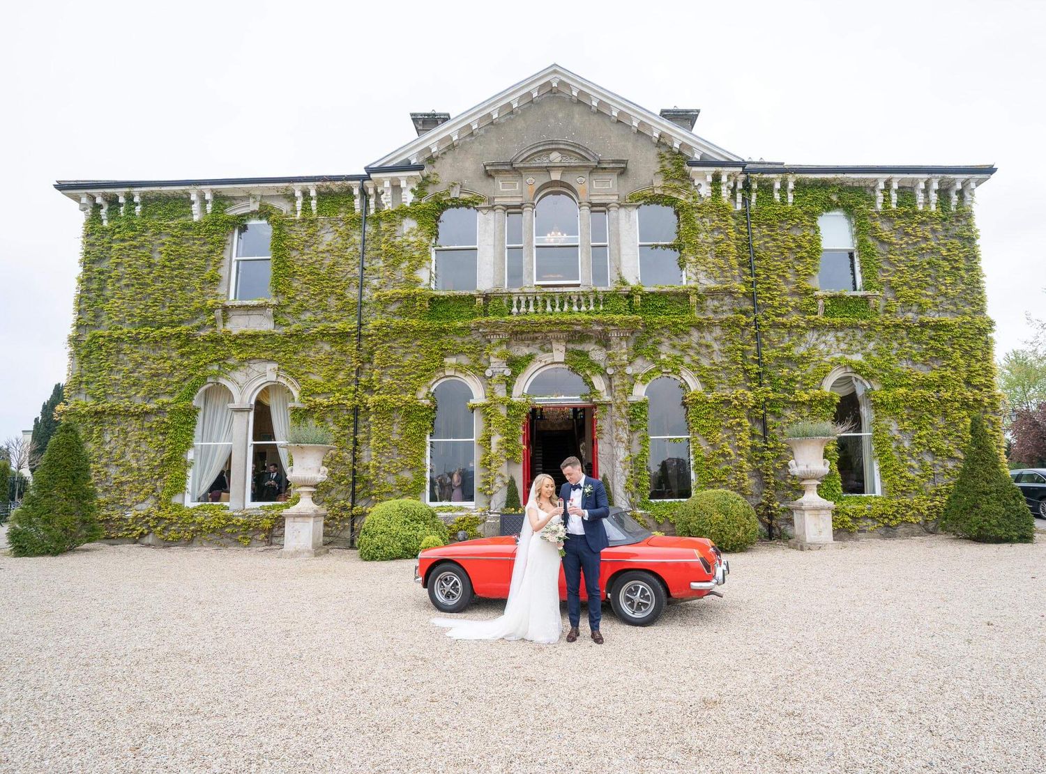 Bride and groom stand beside a red vintage convertible in front Lyrath House photo by AK wedding photography and videography.