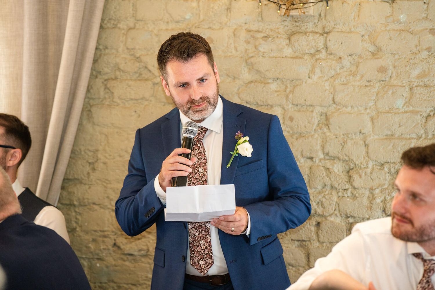 Man in a navy suit with a floral boutonniere speaks into a microphone while holding notes during a wedding speech beside a stone wall.