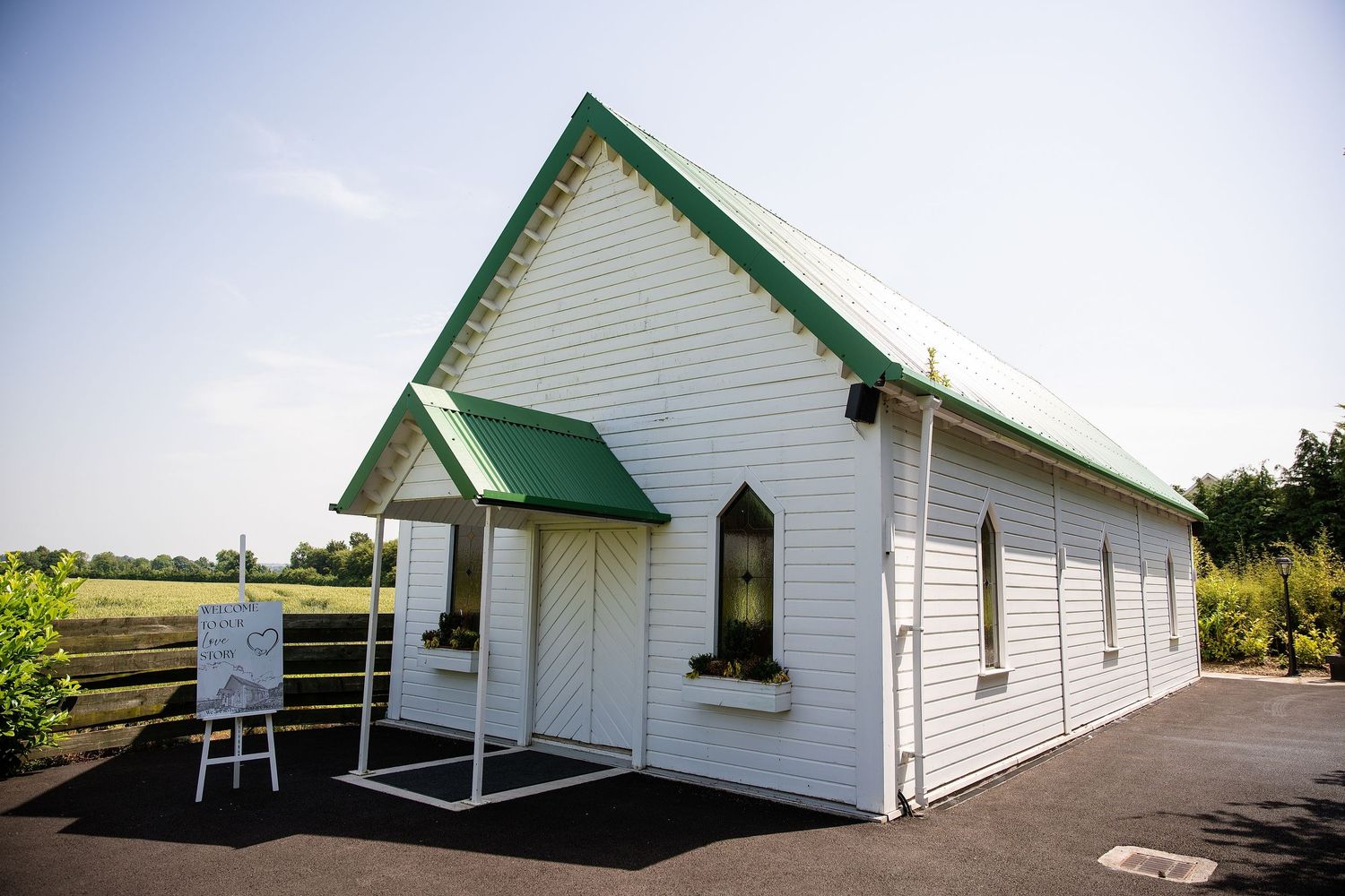 White country chapel with green roof and trim, surrounded by open fields on a sunny day.