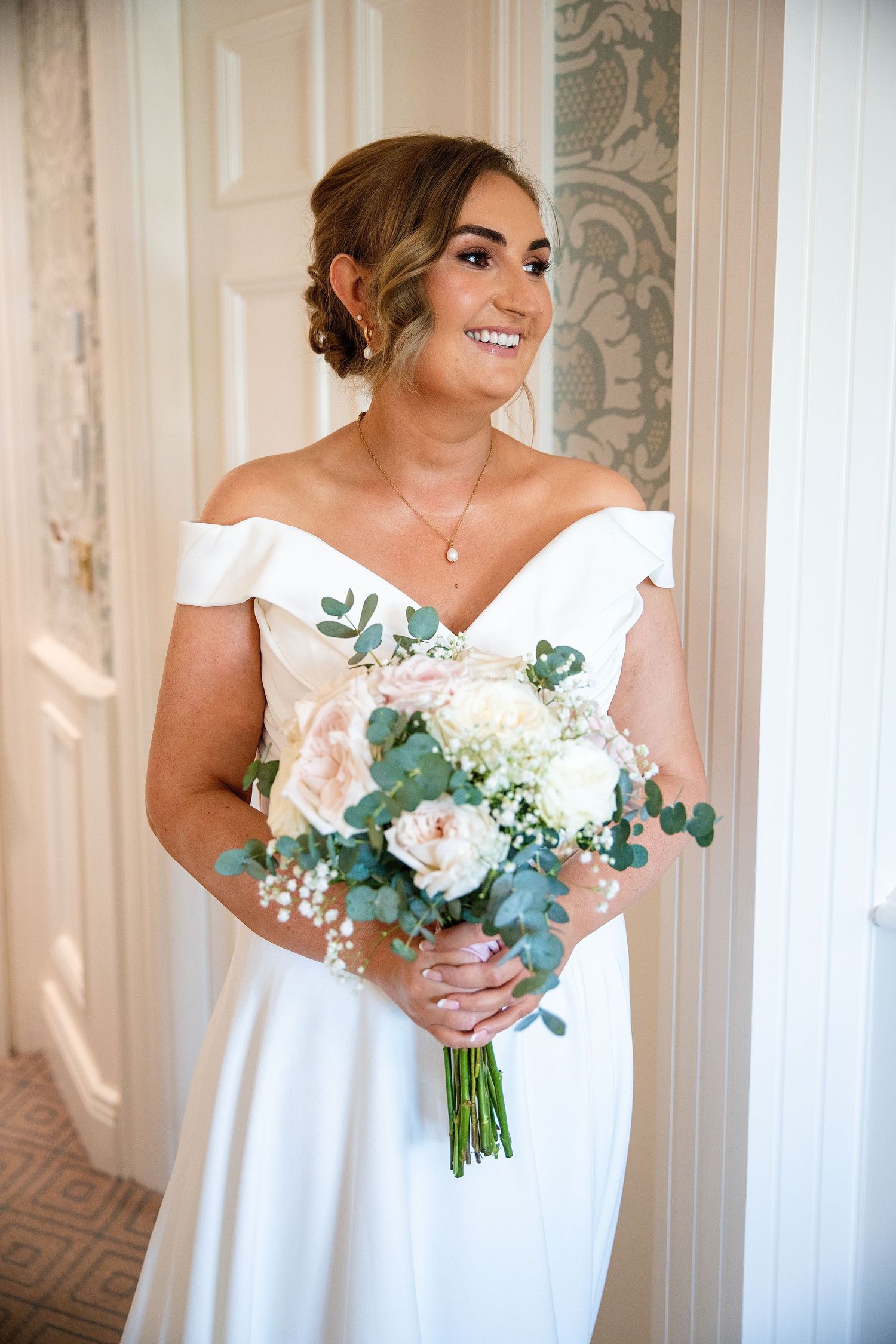 Smiling bride in off-shoulder white gown holding bouquet of white and blush roses with eucalyptus greenery indoors.