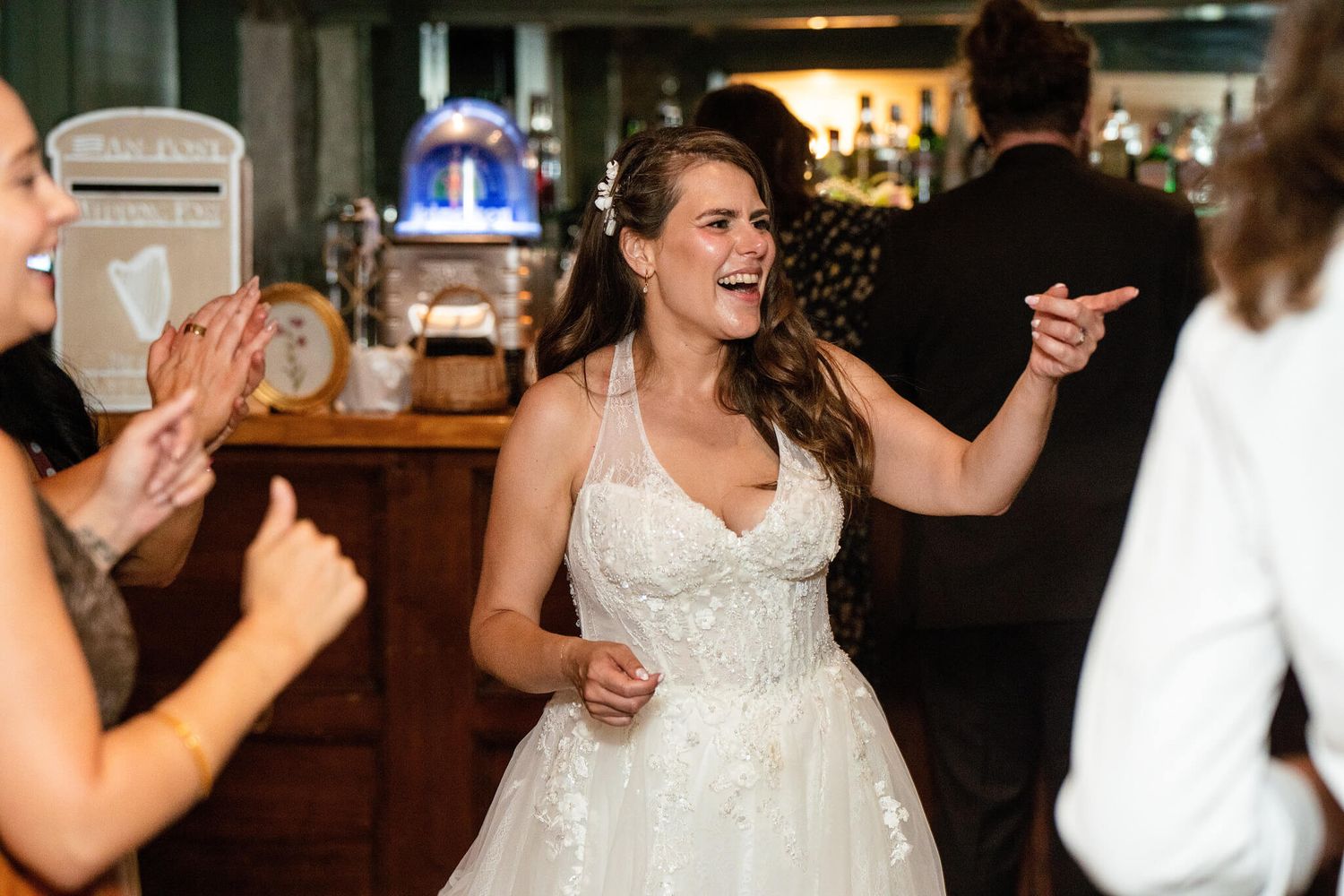 Smiling bride in a white lace wedding gown laughs and dances with guests at a lively reception near a wooden bar.