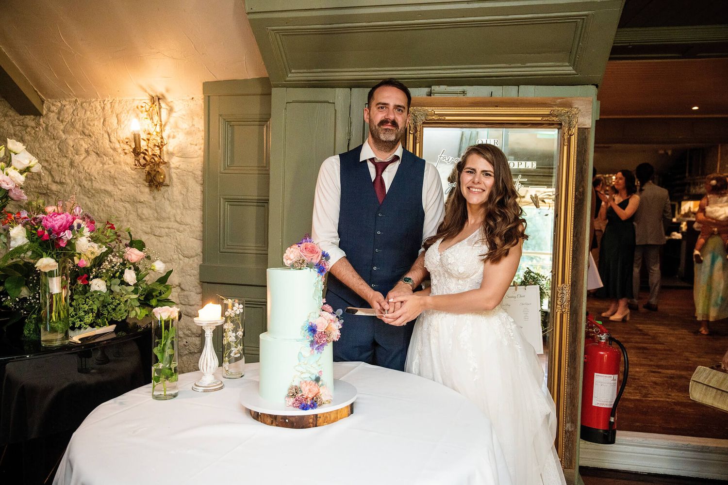 Smiling bride and groom cutting a pastel blue wedding cake decorated with fresh flowers at a rustic reception venue.