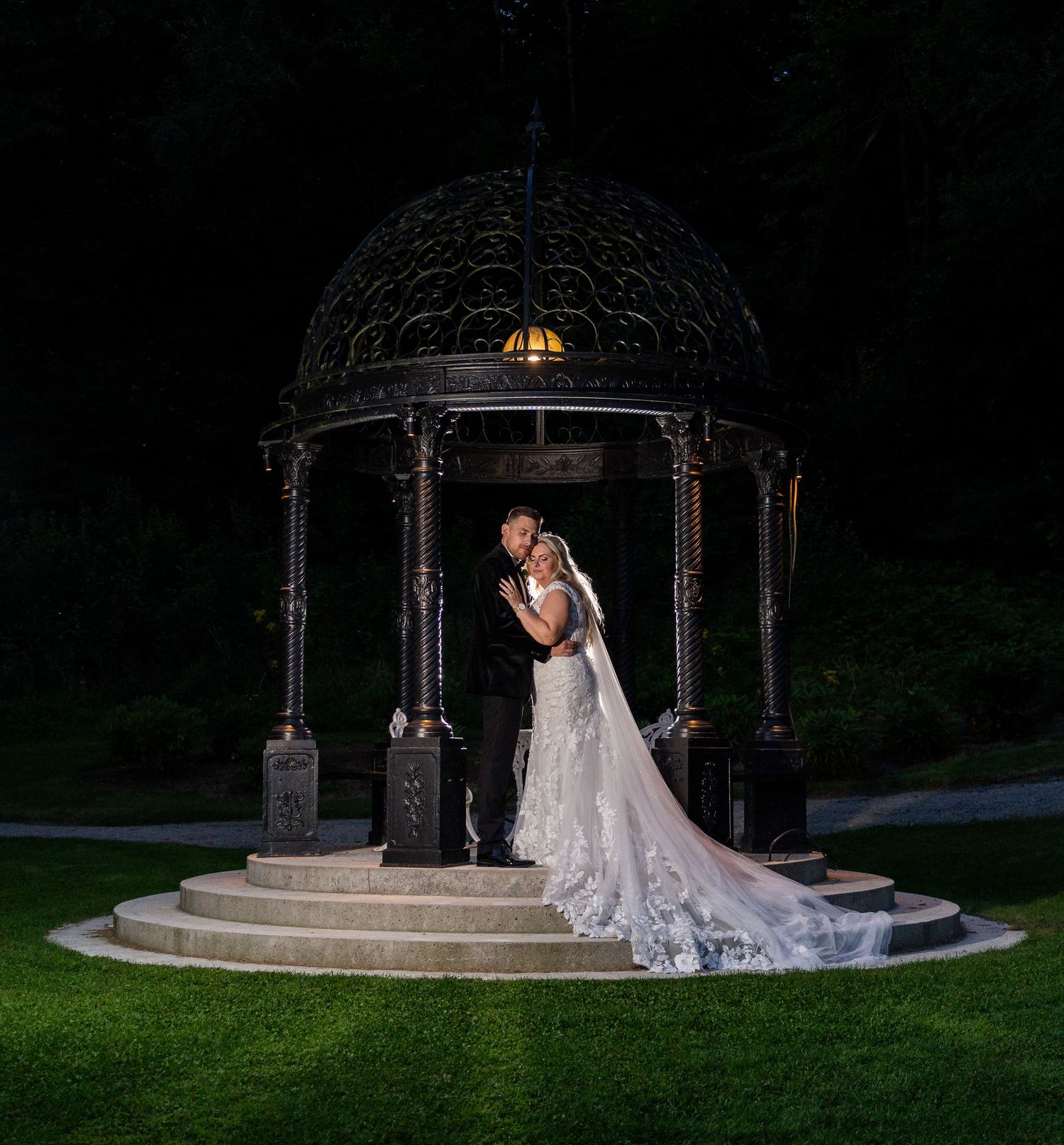A couple shares a romantic moment under an ornate metal gazebo at night, with a dramatic trailing wedding dress.
