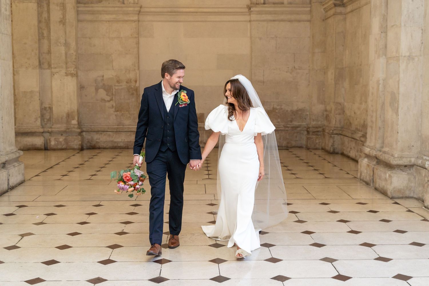 Bride and groom walk hand-in-hand in elegant stone hall, bride in white gown with veil, groom in navy suit.