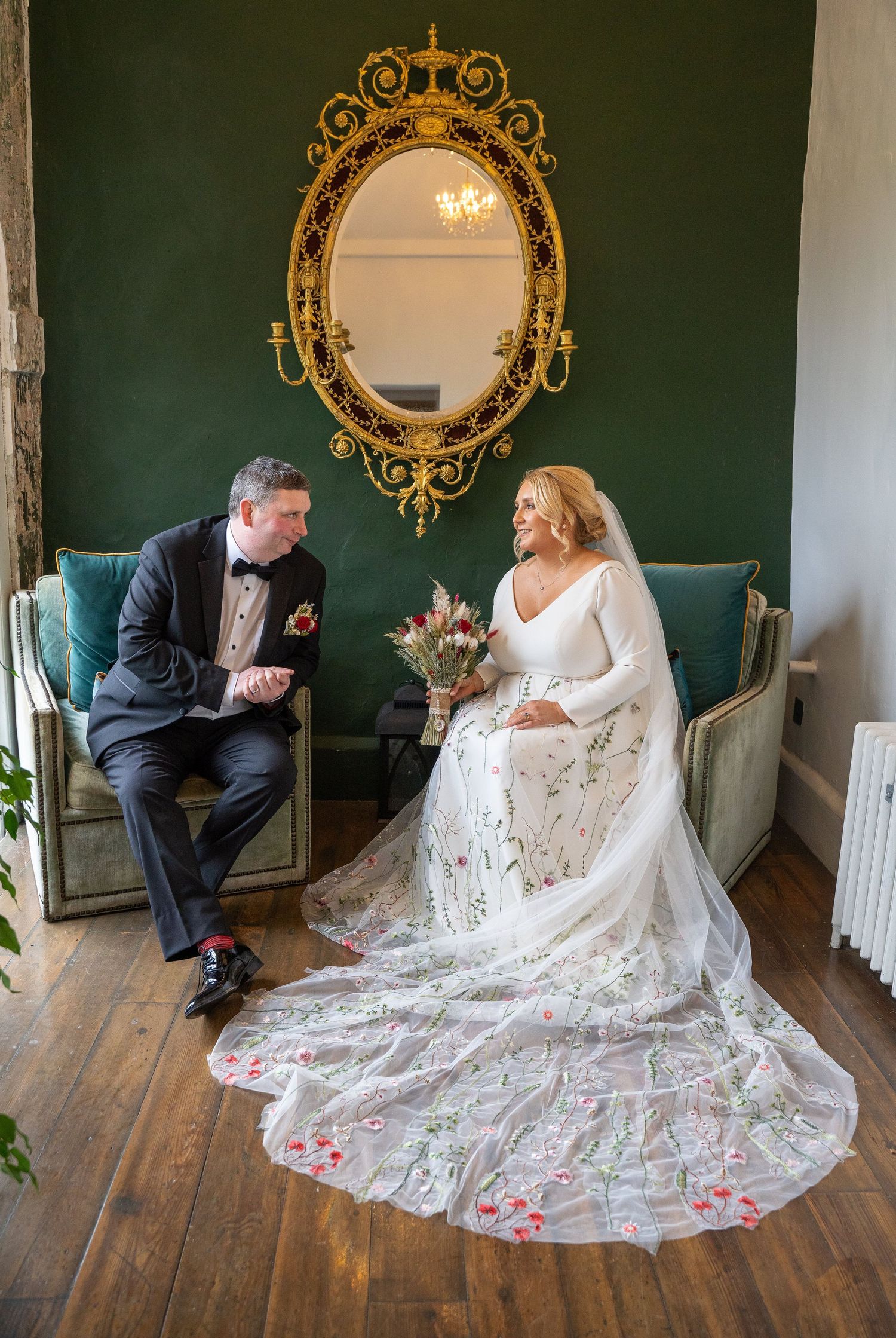 Bride in floral embroidered wedding gown with long veil sits beside groom in tuxedo before ornate gold mirrors at Bellinter house.