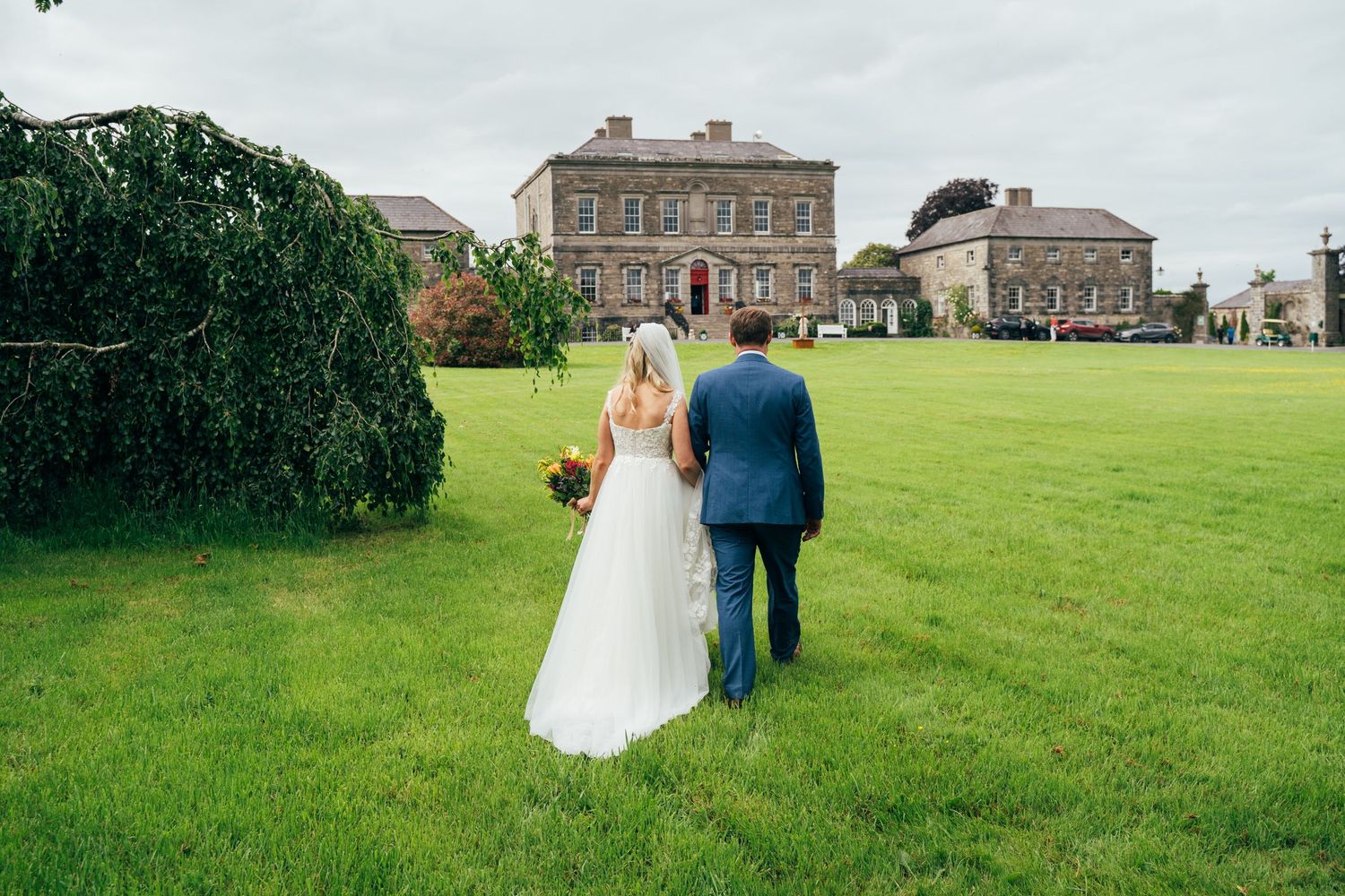 Bride and groom walking hand in hand across lush green lawn toward elegant stone country house wedding venue at Bellinter house.