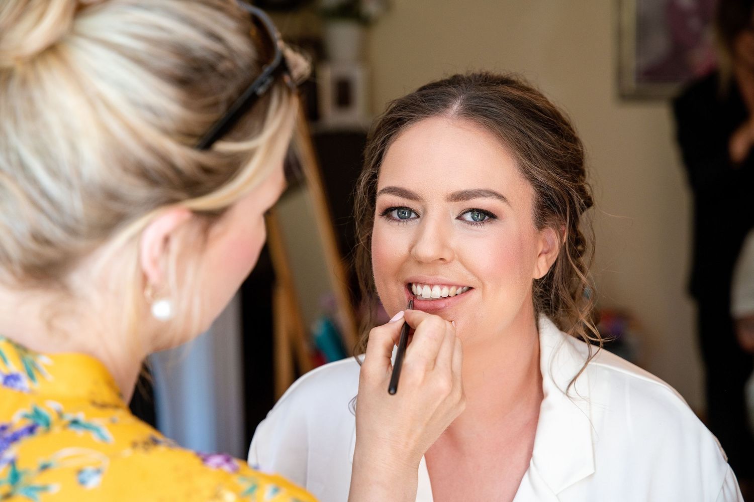 Makeup artist applying lipstick to a smiling bride in a white robe during bridal preparation.