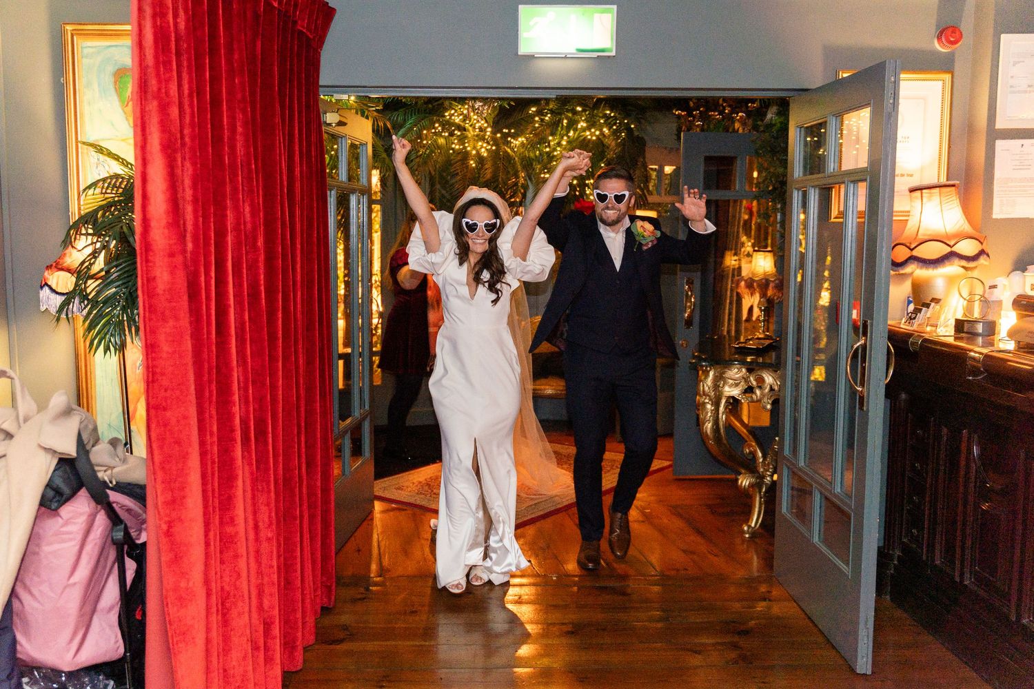 Bride and groom making a grand entrance at their wedding reception, wearing sunglasses and raising their arms in celebration.