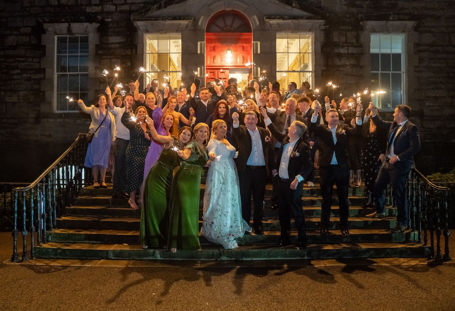 Wedding party celebrating on steps of Bellinter House at night, holding sparklers, guests in formal attire.