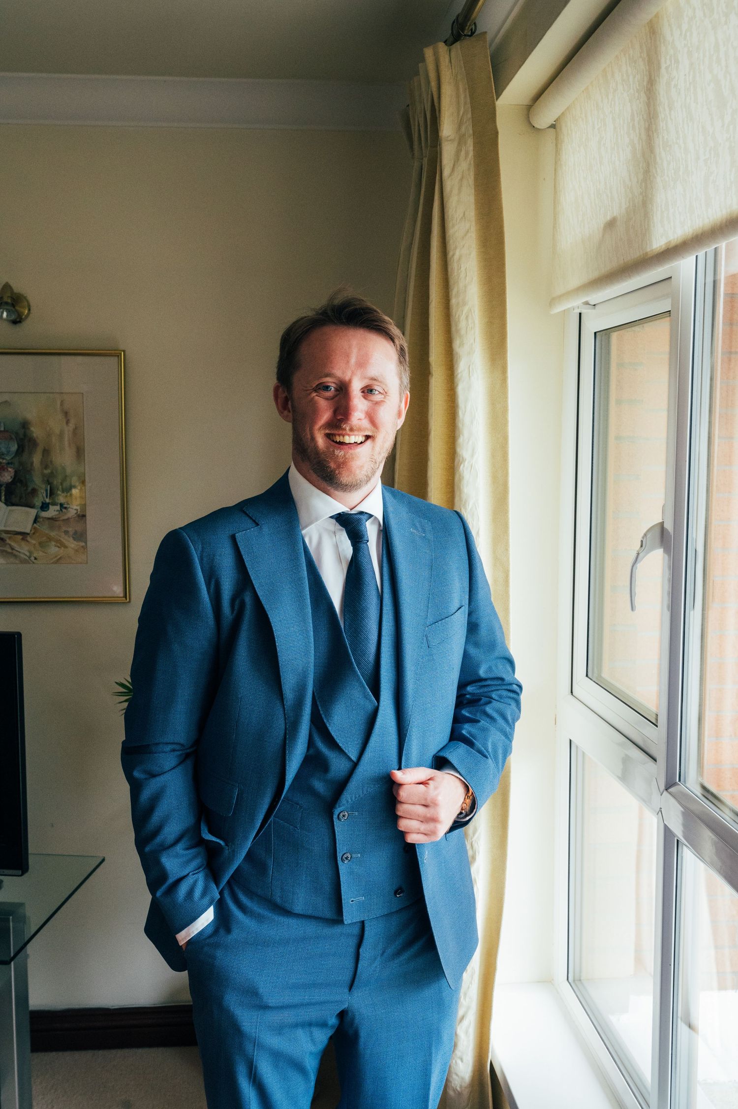 Smiling man in a blue three-piece suit standing by a window in a well-lit room.