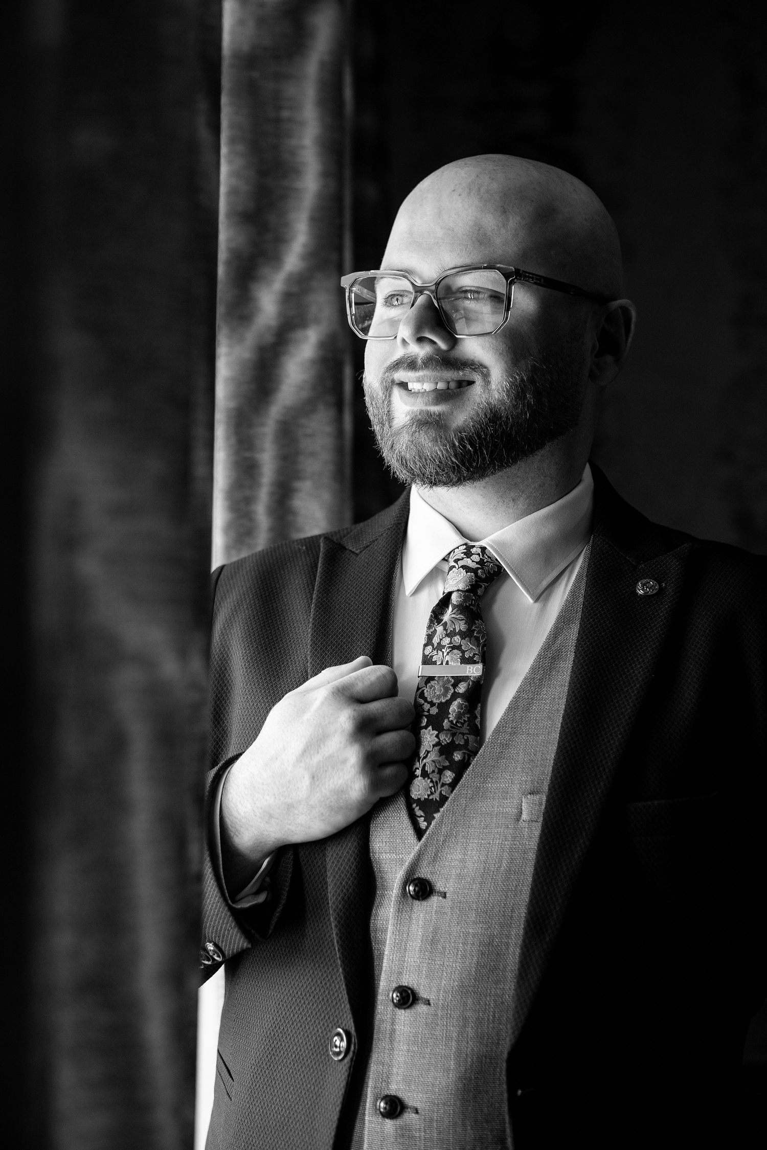 Smiling bald man with beard and glasses wearing a suit, vest, and floral tie in a black and white portrait.
