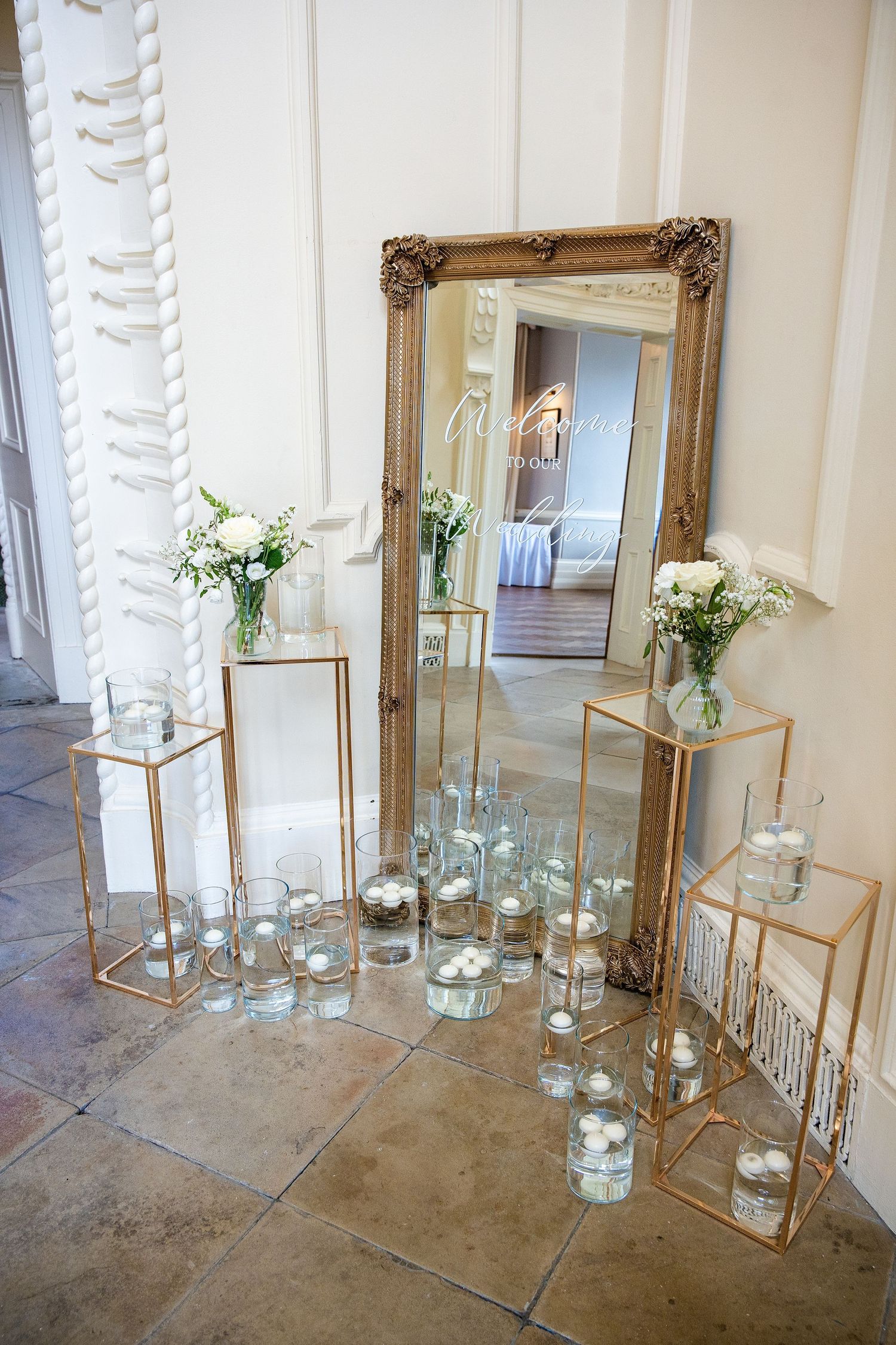Elegant wedding decor with gold-framed mirror, glass terrariums, white floral arrangements, and candles on gold stands.