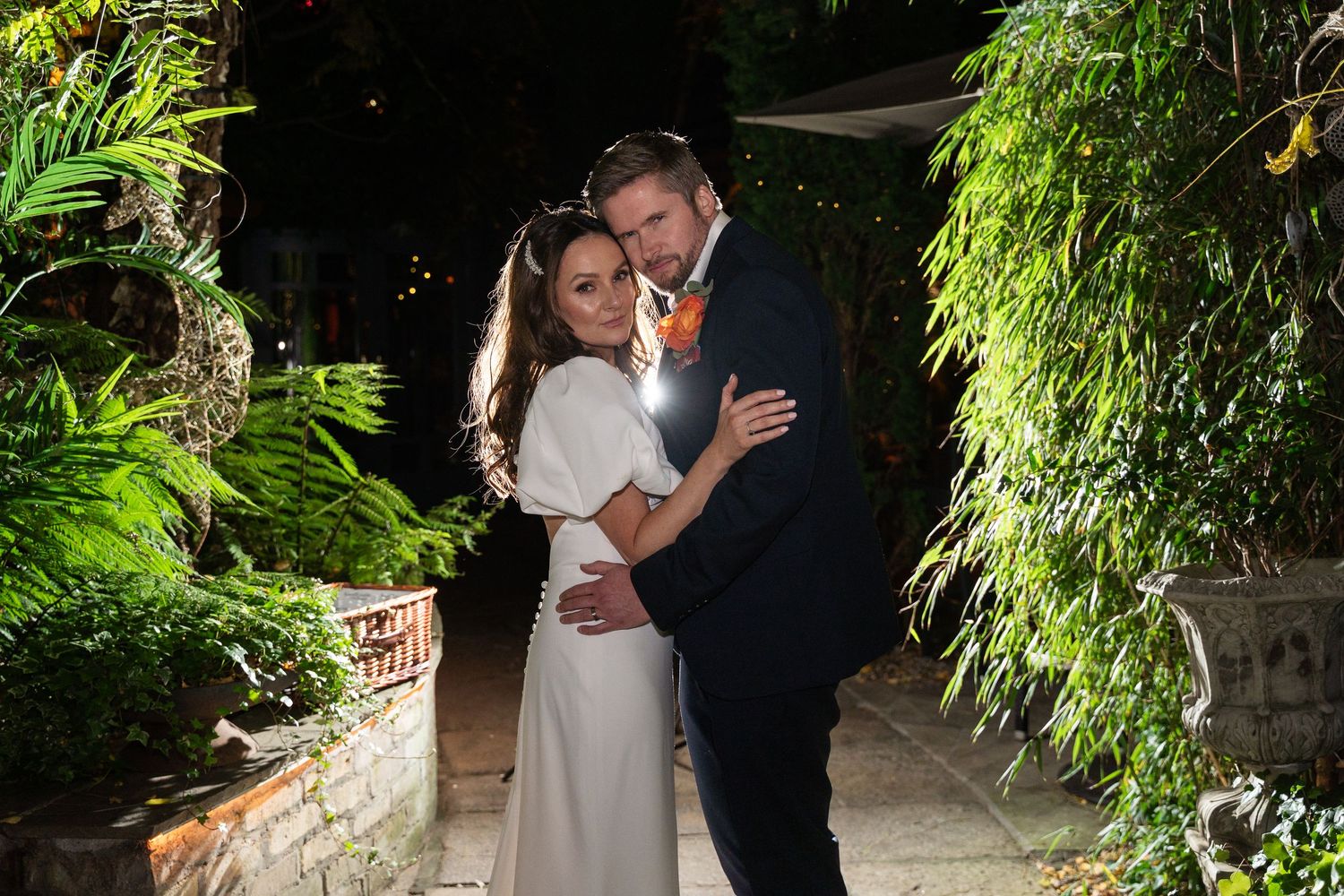 Bride and groom embracing outdoors at night surrounded by lush greenery at a romantic garden wedding venue.