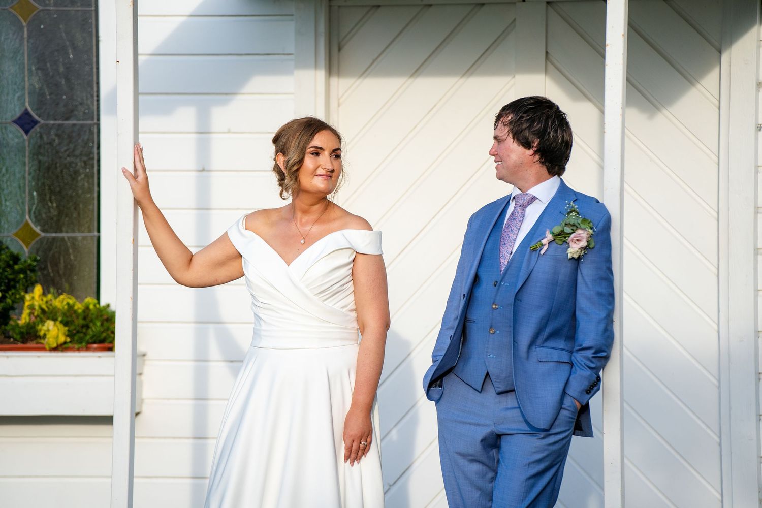Bride in white off-shoulder gown and groom in blue suit share a joyful moment outside white wooden doors.
