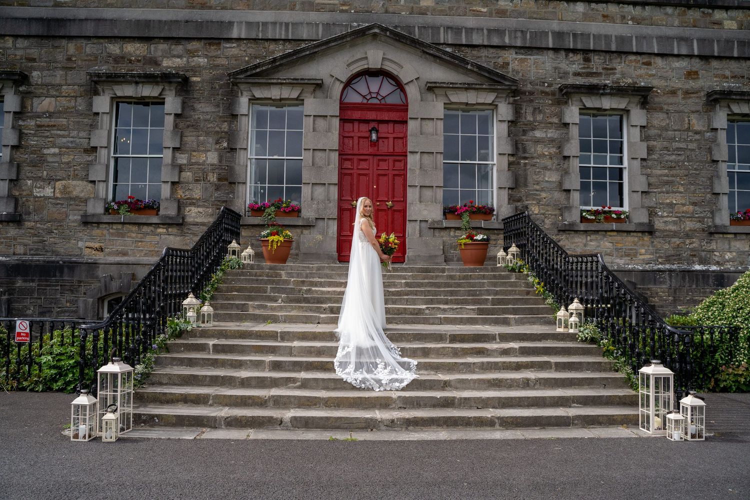 Bride in white gown and veil stands on stone steps before a grand building with a red door and lanterns at Bellinter house.