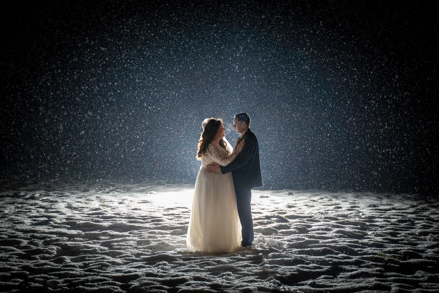Bride and groom share a romantic embrace in falling snow at night, her white wedding gown glowing against the dark winter backdrop.