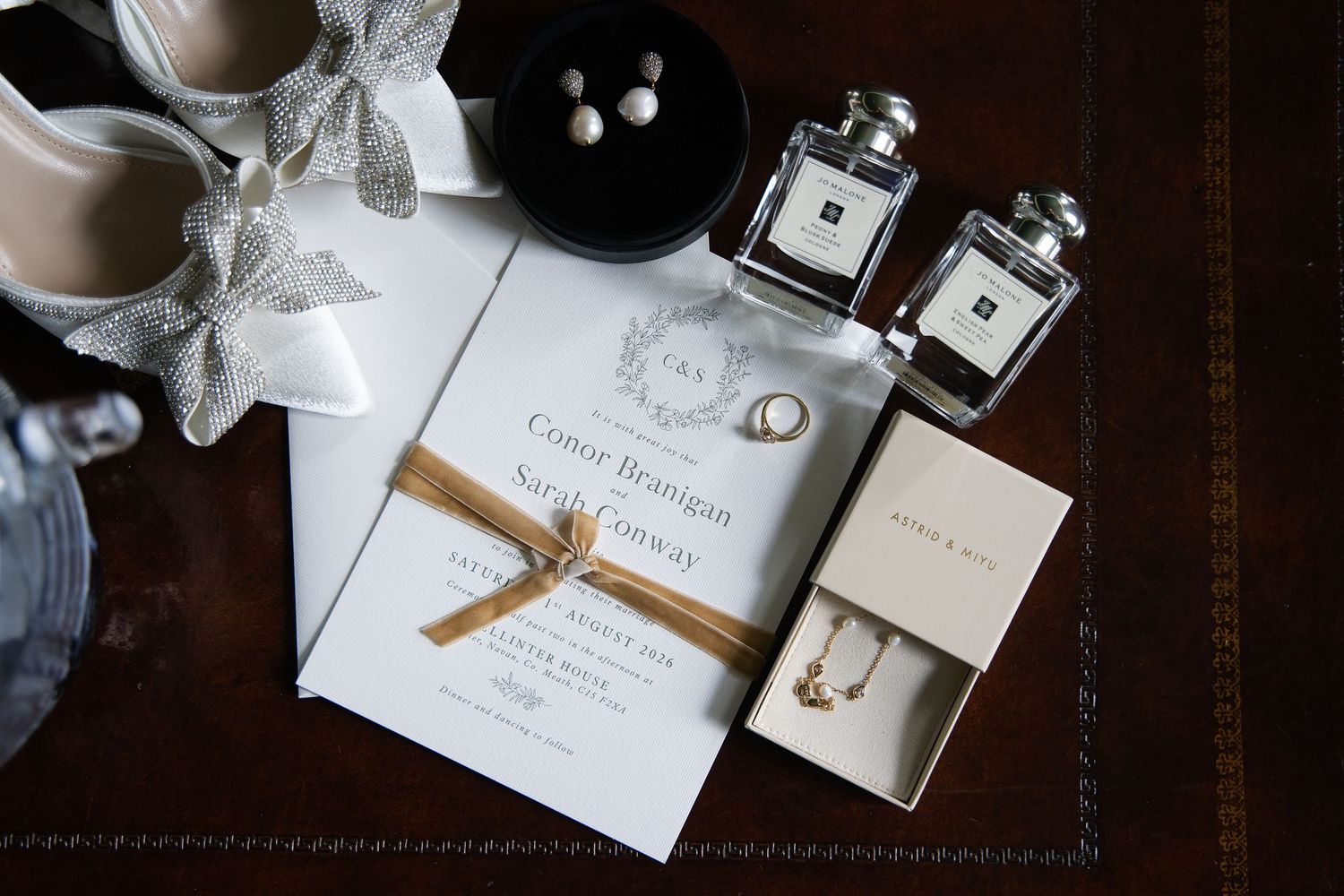 Flat lay of wedding invitation, Jo Malone perfume, jewelry, and accessories on wood surface.