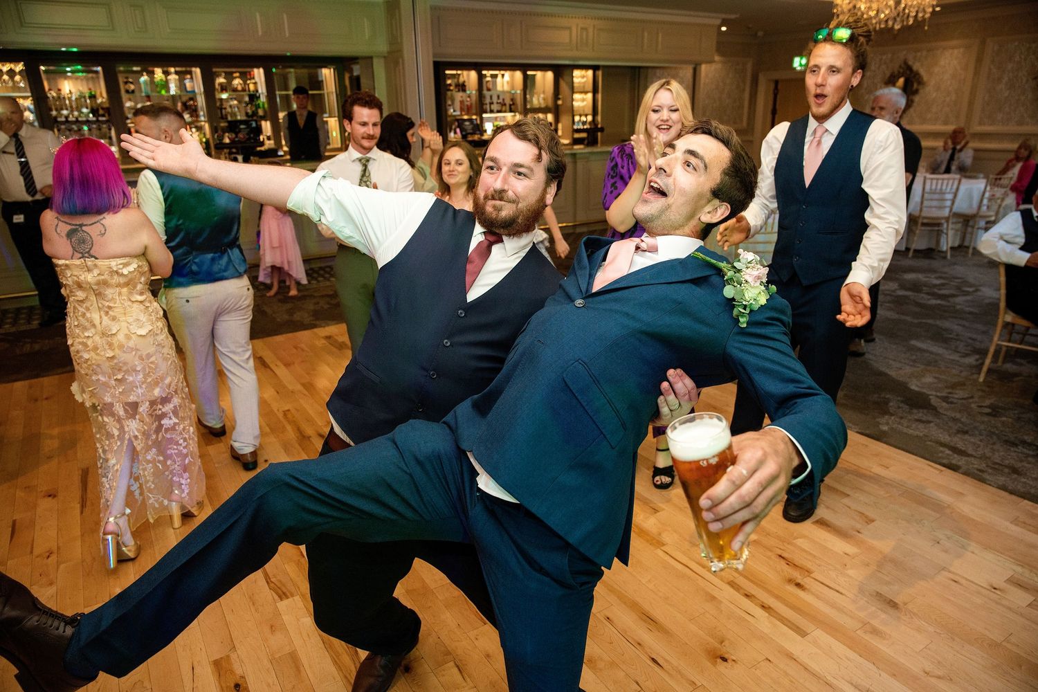 Two groomsmen dancing energetically at a wedding reception, one holding a beer glass on a wooden dance floor.