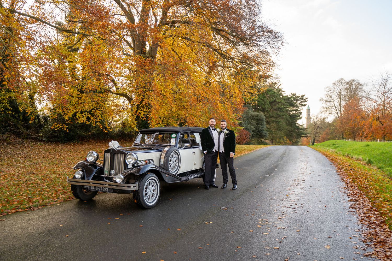 Married couple in formal suits stand beside a vintage car in the Phoenix park Dublin photo by AK wedding photography and videography.