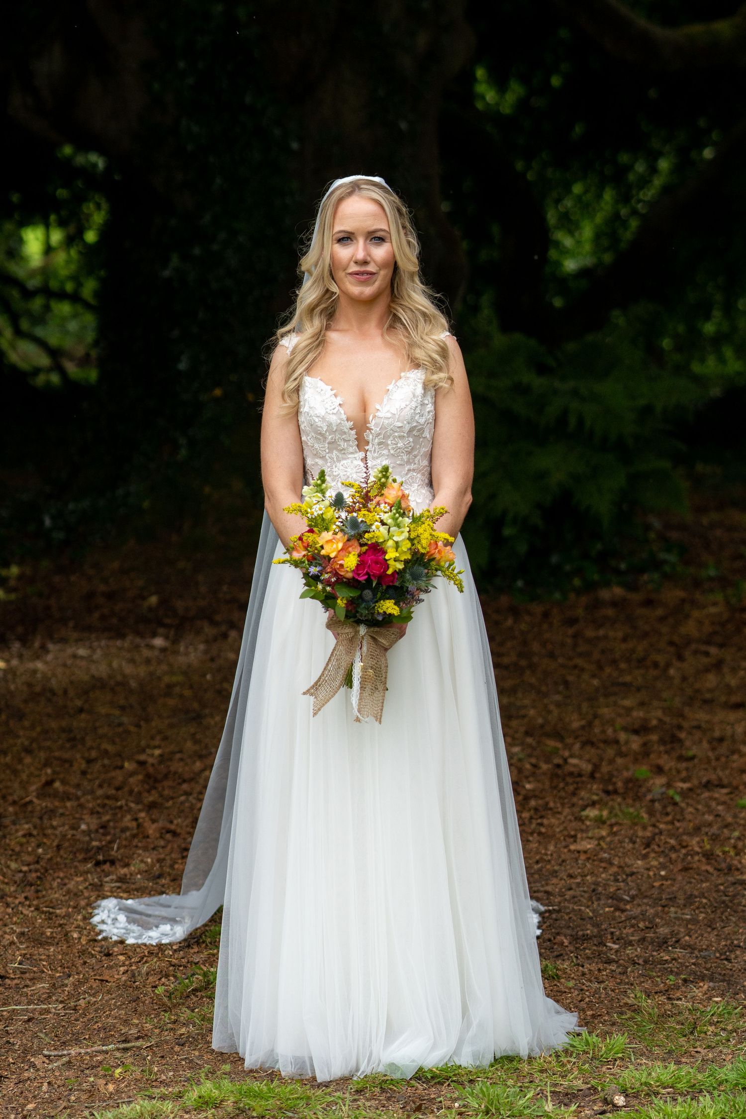 Bride in a white tulle gown holding a colorful bouquet of flowers, standing outdoors among trees at Bellinter house.
