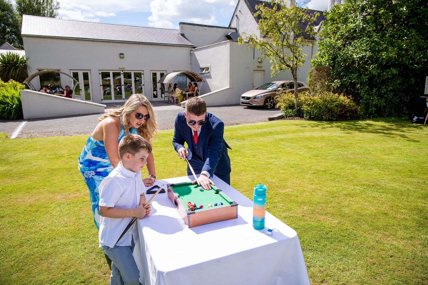 Two guests and a child play tabletop pool on a white draped table outdoors at a sunny wedding reception.
