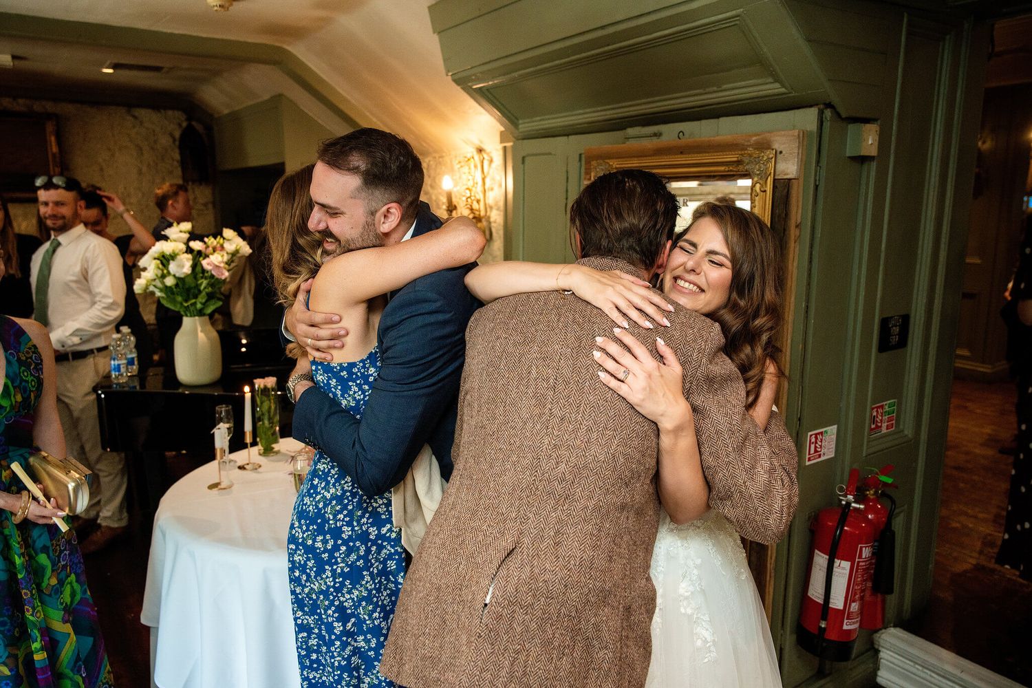Wedding guests share a warm group hug beside a table with flowers and candles in a cozy, rustic indoor venue.