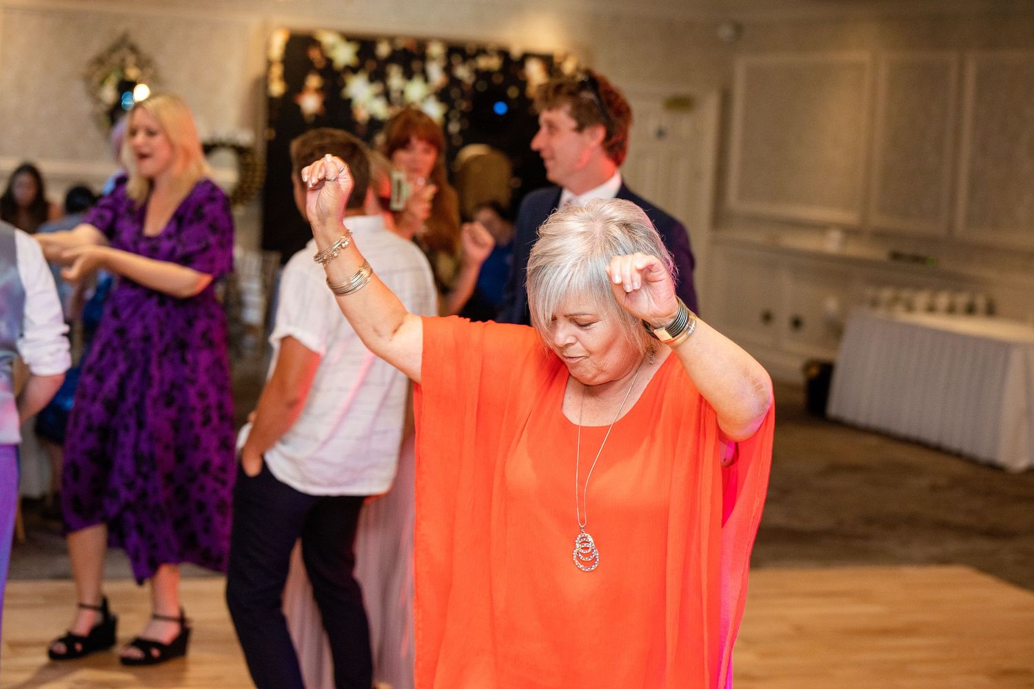 Woman in orange dress dancing joyfully at a wedding reception with guests celebrating in the background.