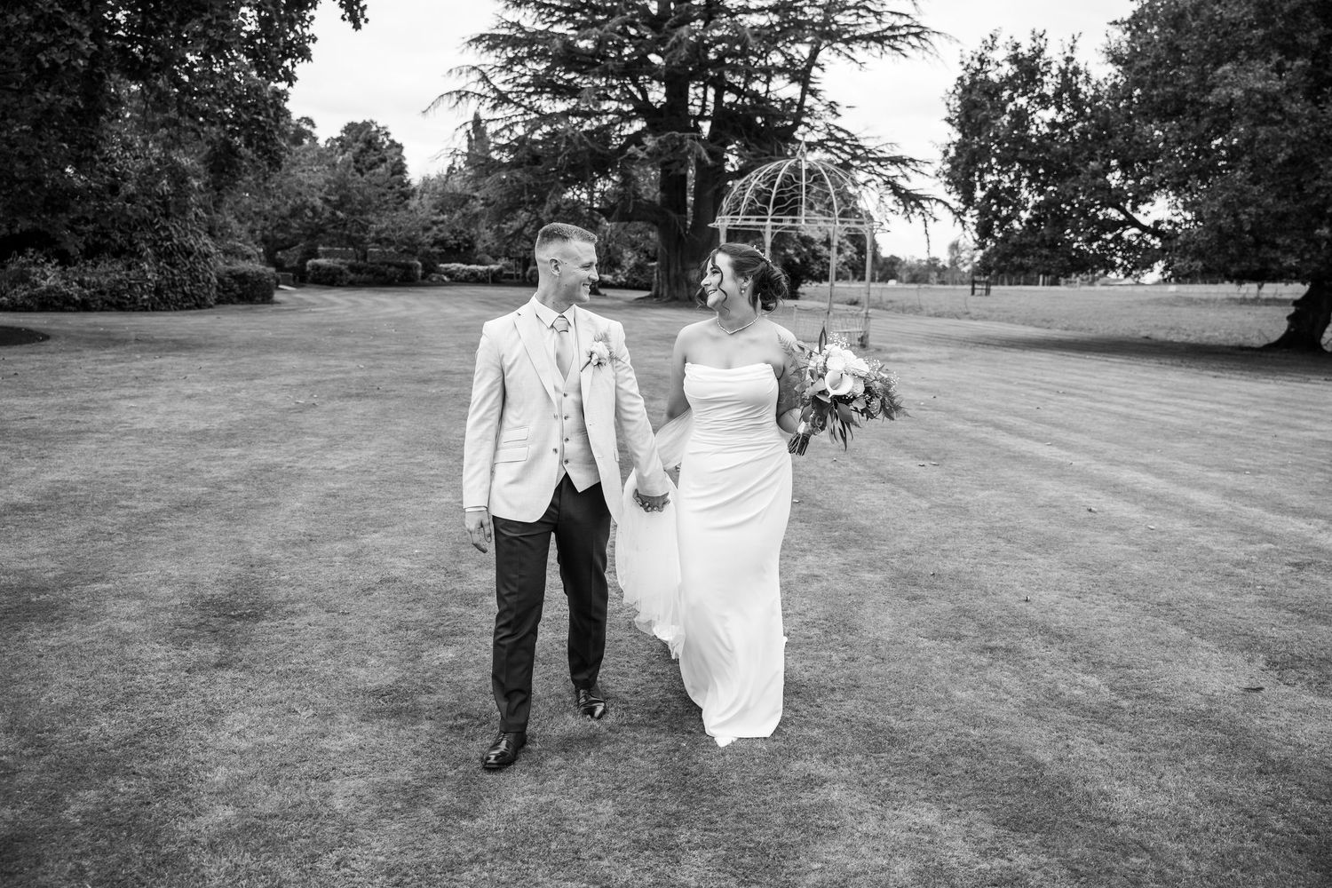 Black and white photo of a bride and groom holding hands walking across a lawn on their wedding day.