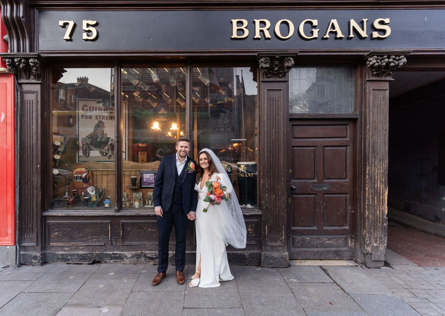 Bride and groom posing outside Brogans pub at number 75, smiling on their wedding day.