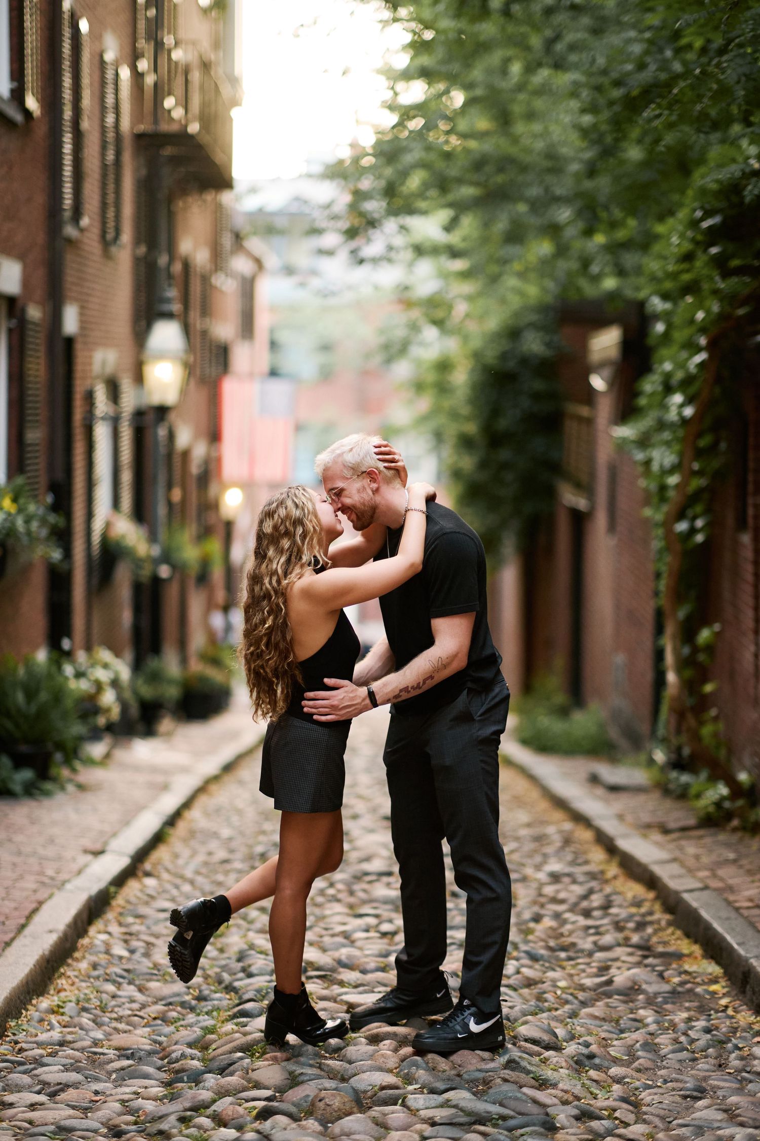 A couple shares a romantic kiss on a cobblestone street in Boston's Acorn Street, both dressed in black.