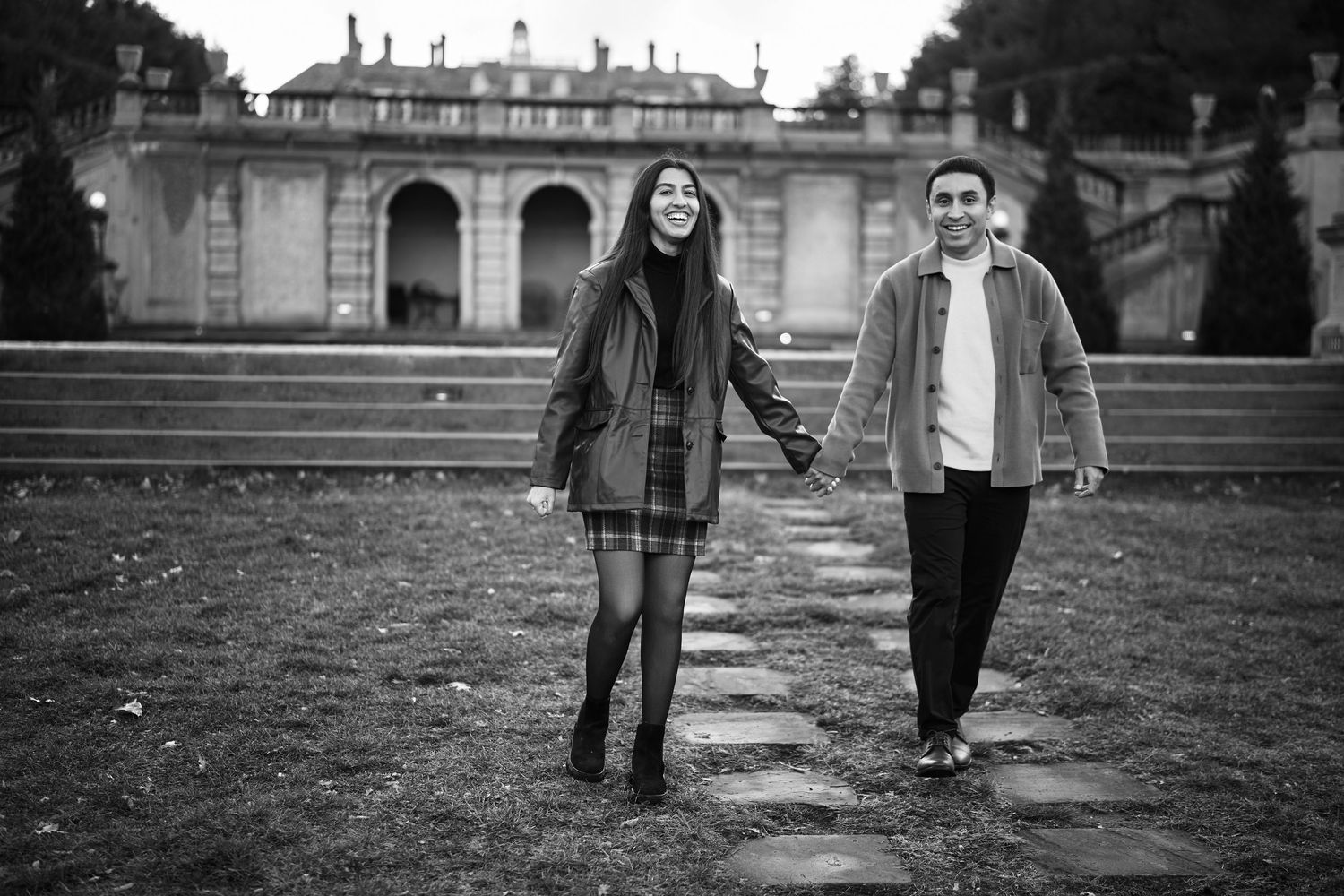 Black and white photo of a smiling couple holding hands walking in a park with a grand building behind them.
