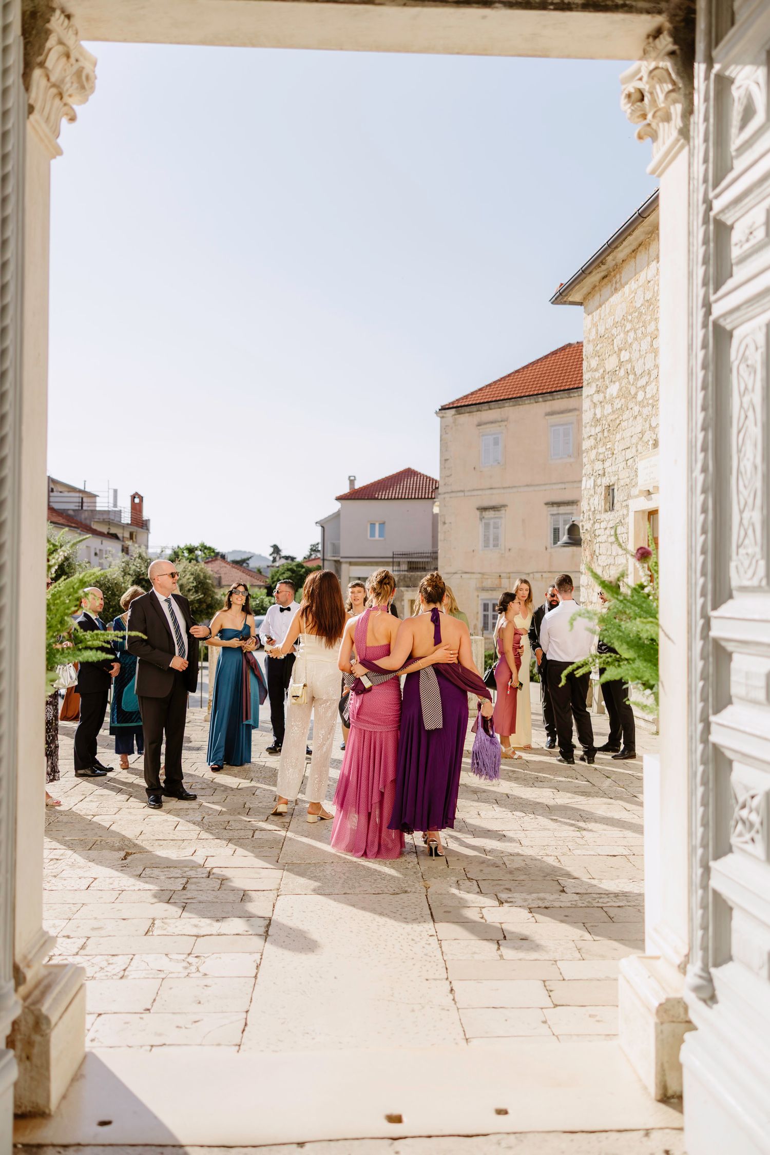 Wedding guests mingling on a sunny stone terrace outside a Mediterranean-style building.