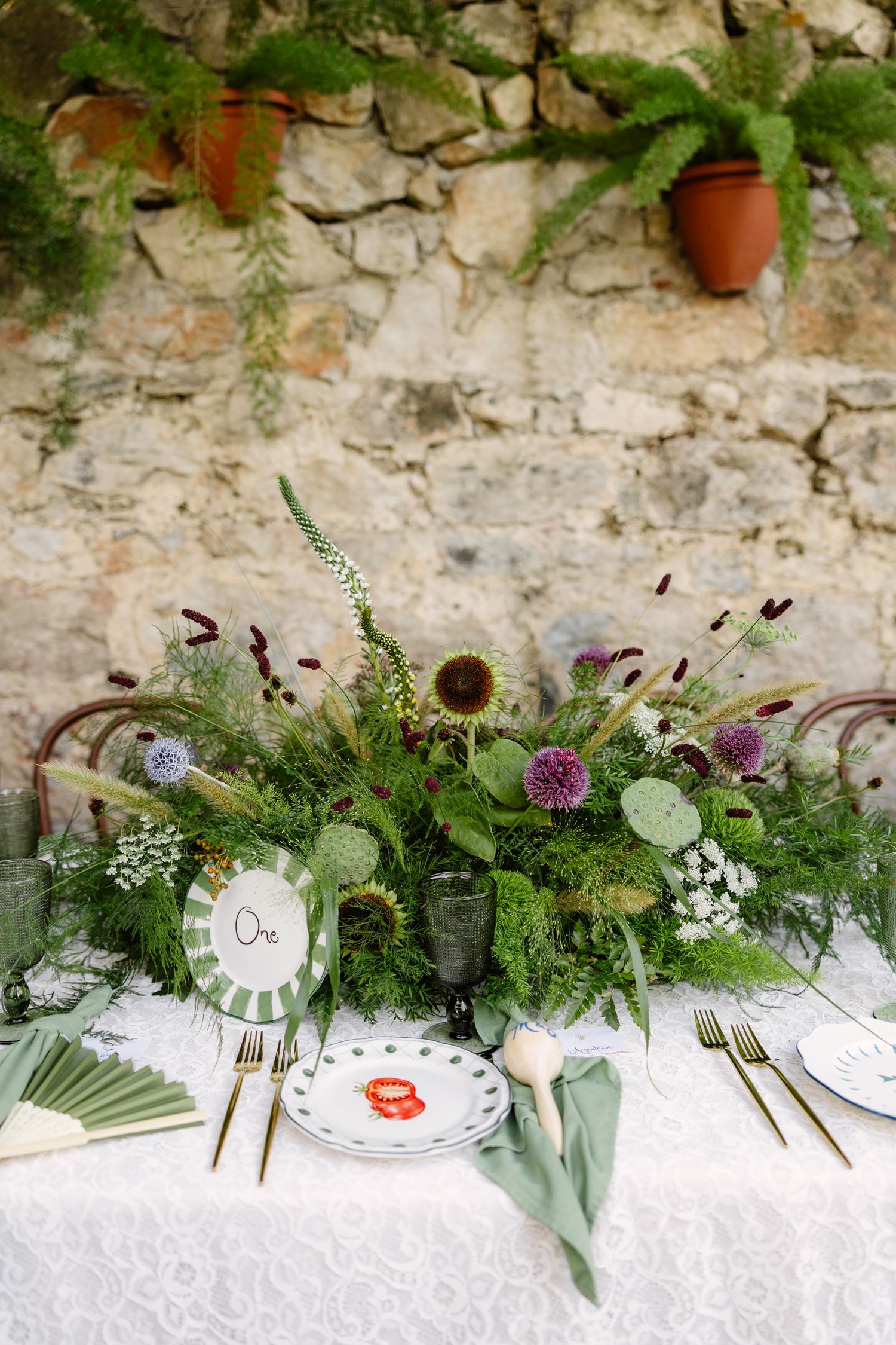 Wedding table decor with white linens and natural greenery against a textured stone wall.