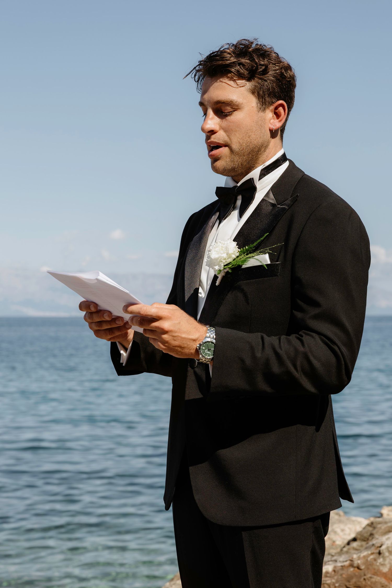 Groom in classic black tuxedo reading from paper against ocean backdrop.