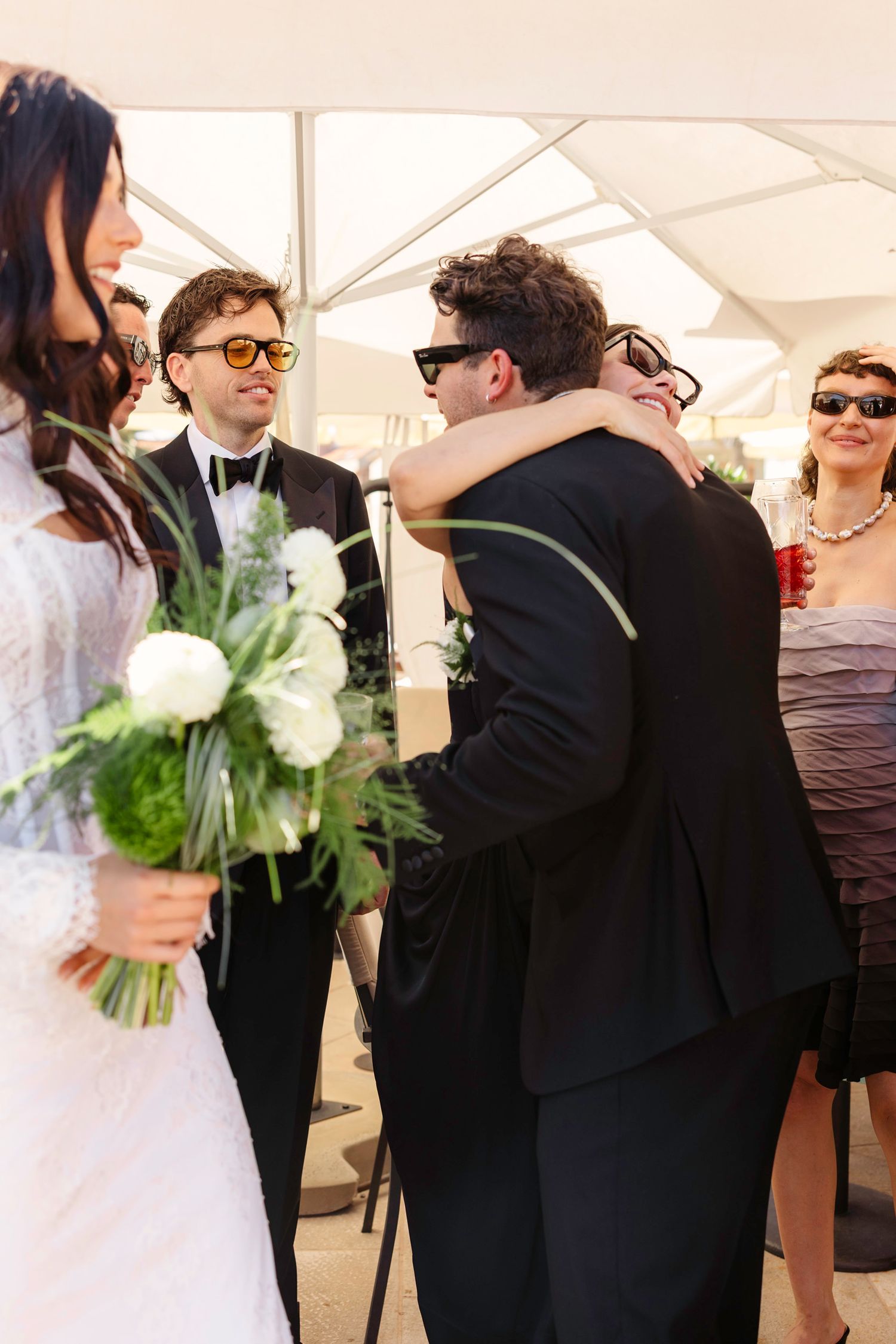 Wedding guests embrace during an intimate ceremony while the bride holds a white and green bouquet.