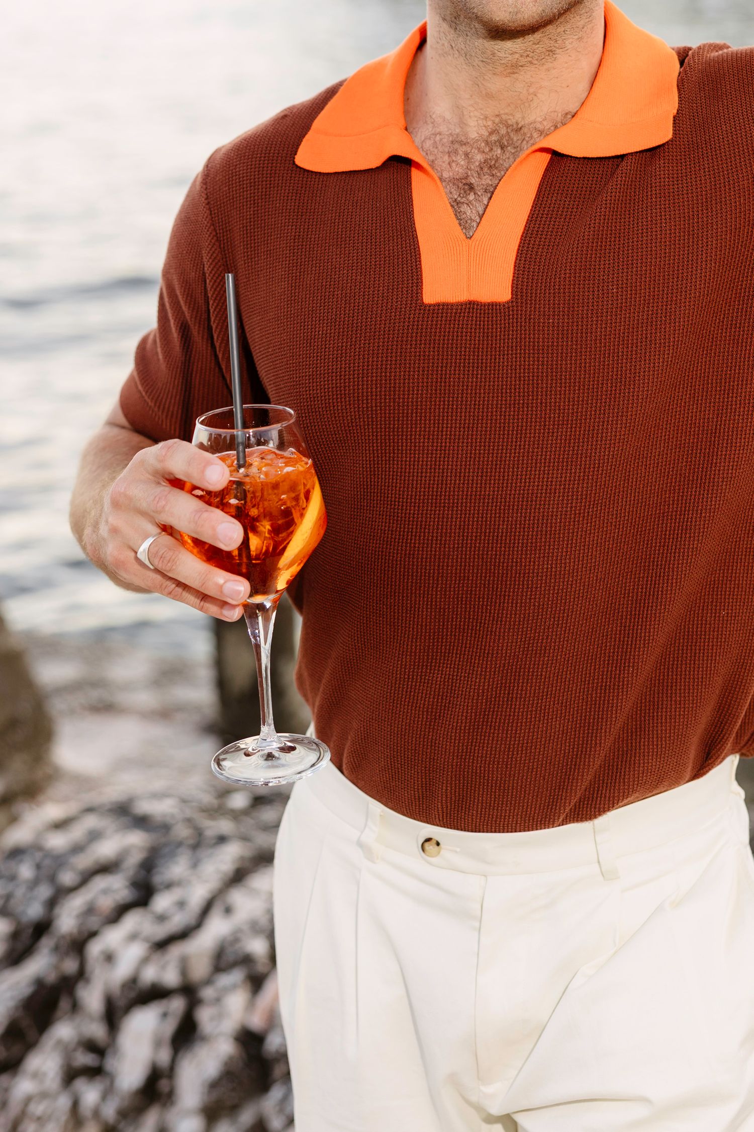 Close-up of person wearing orange-collared brown shirt holding cocktail glass by the ocean.