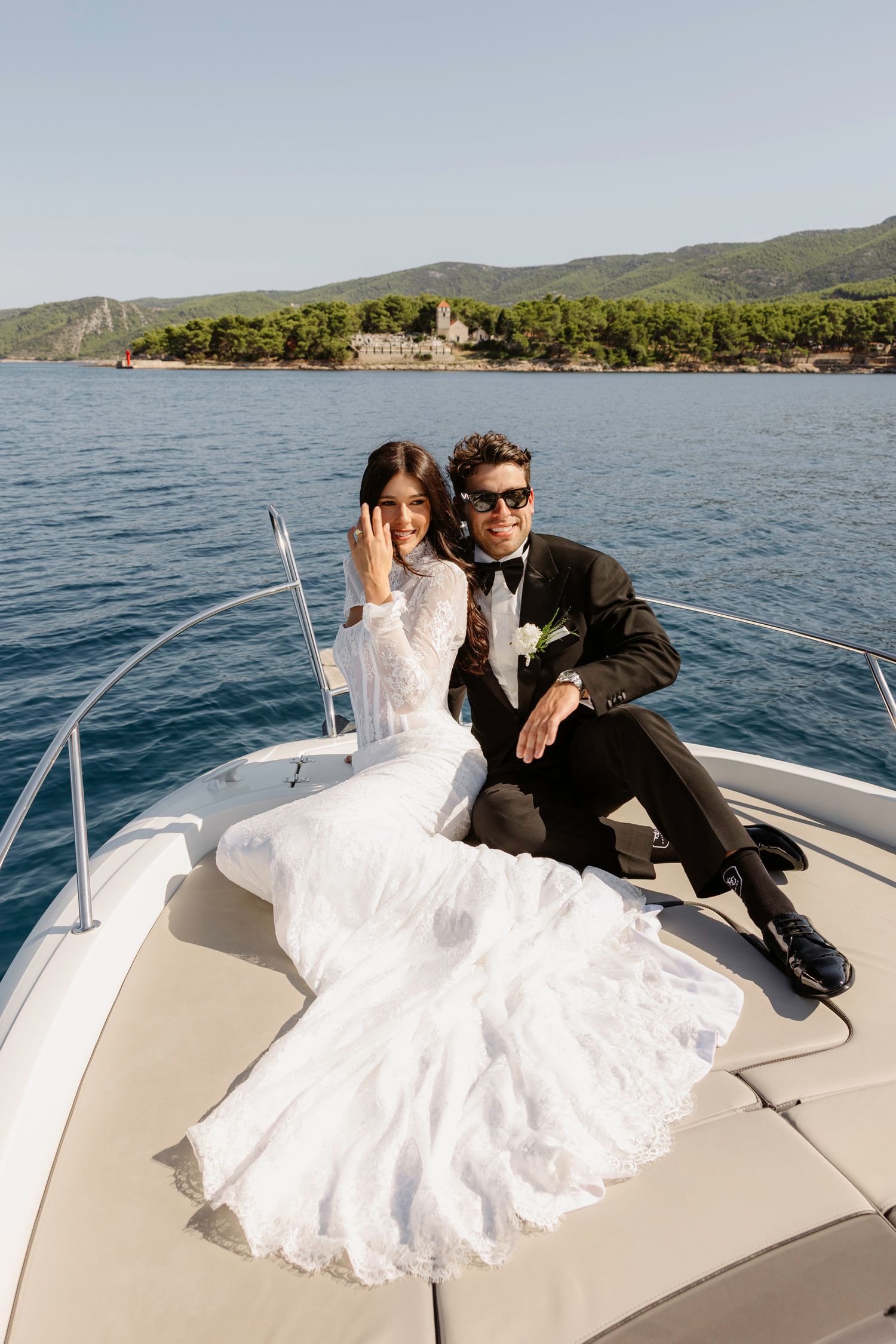 Newlyweds sitting together on bow of boat with coastal scenery behind them.