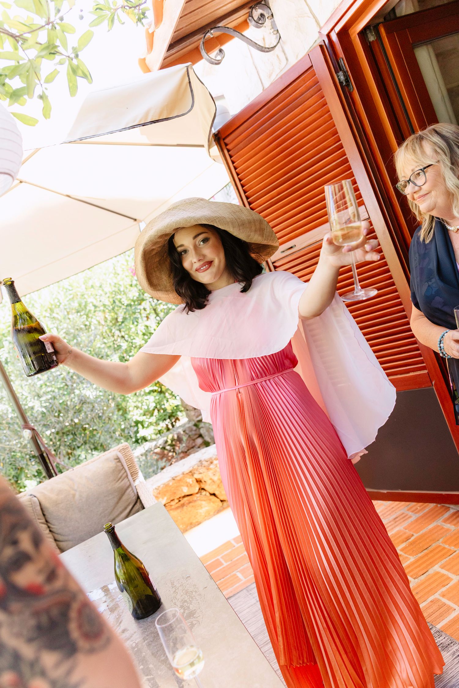 Woman in coral dress and sun hat enjoying champagne on outdoor patio.
