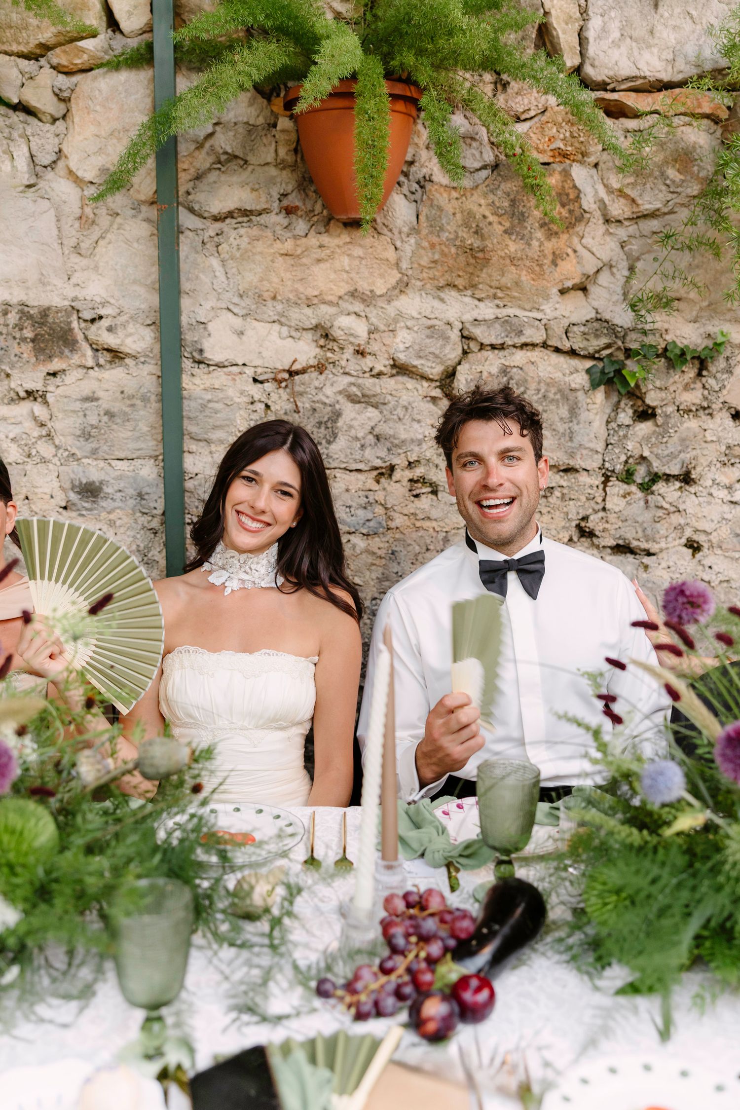Wedding couple smiles and laughs while sitting at a rustic stone wall decorated with greenery and flowers. Hvar, Croatia