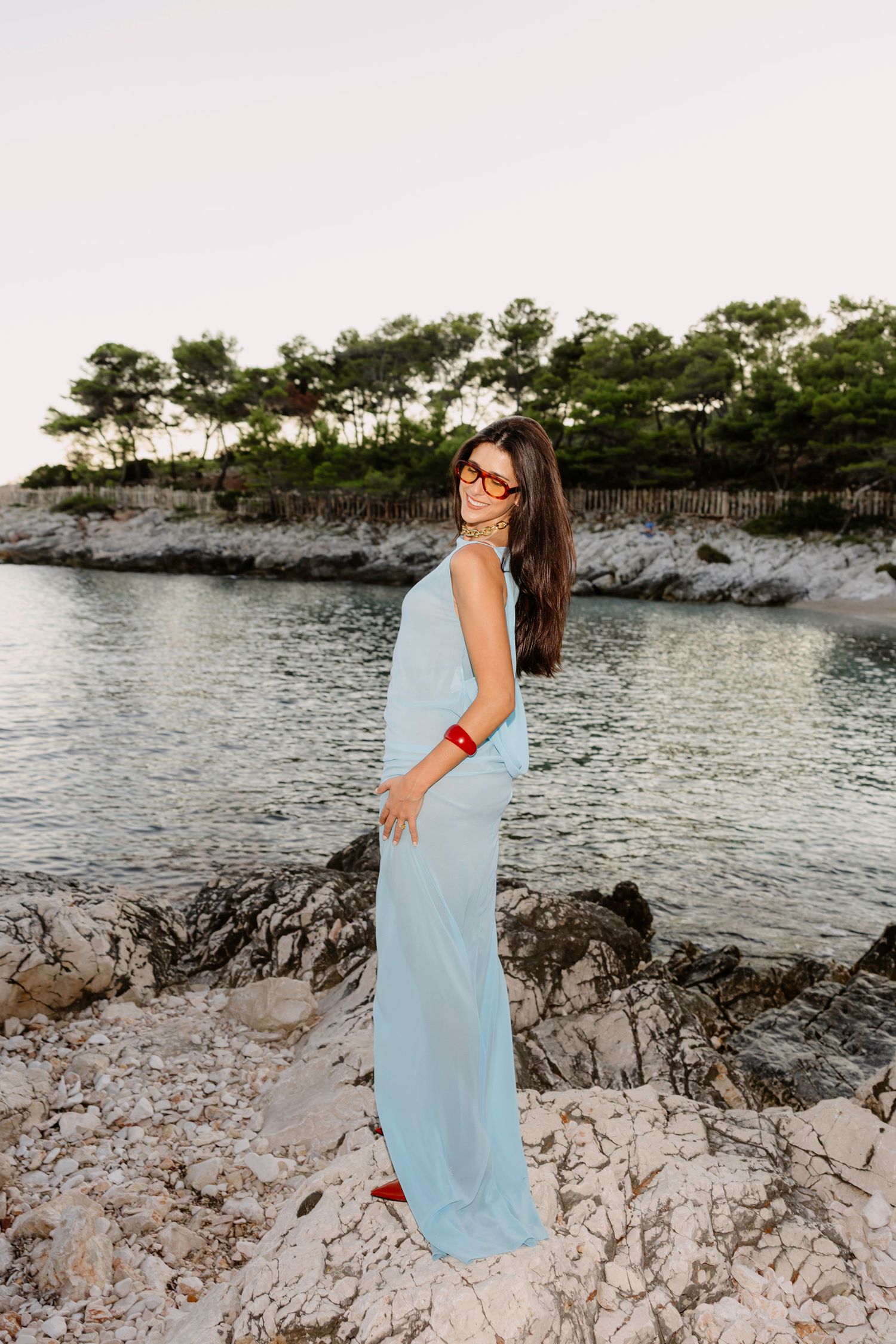A woman in flowing blue dress poses gracefully against a coastal landscape at sunset.