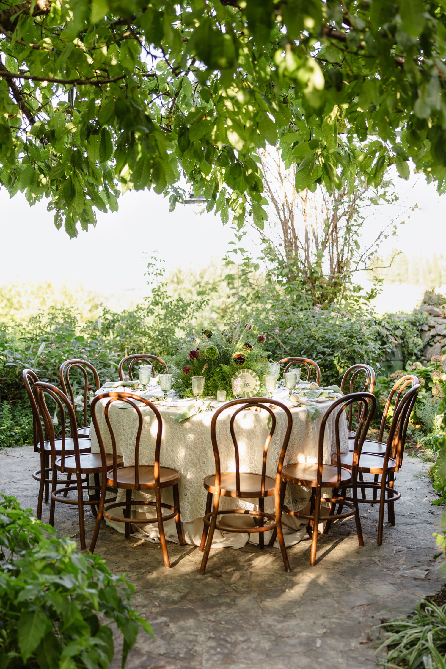 Garden reception setup with wooden chairs and white tablecloths under leafy green canopy.