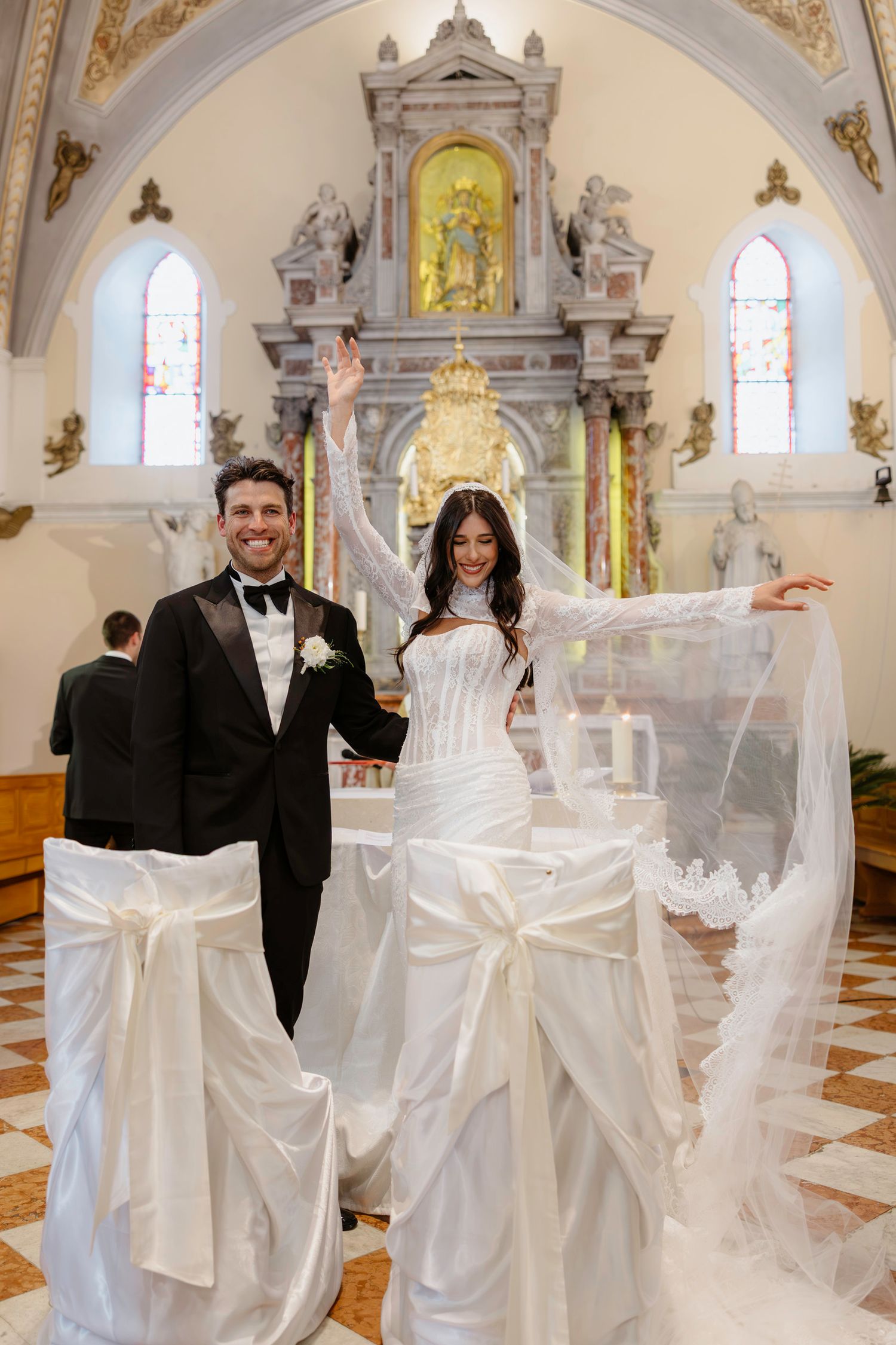 Newlyweds celebrate in front of ornate church altar with decorated white chairs.