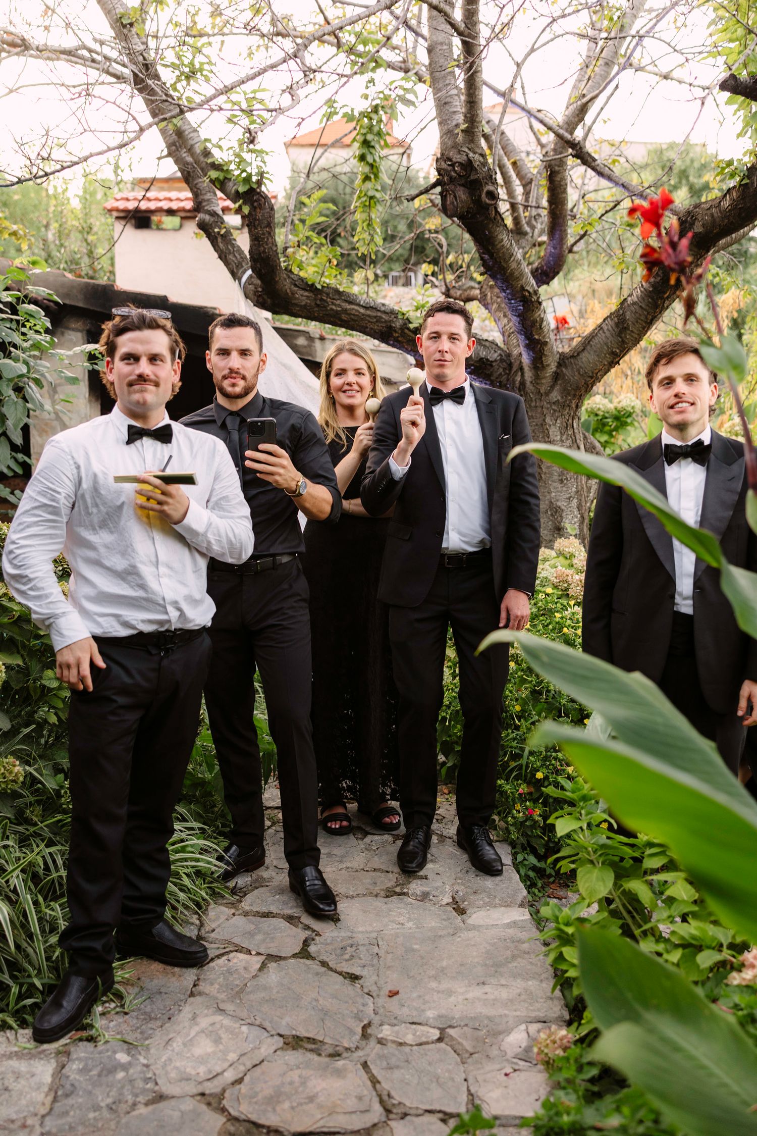 Wedding guests in formal attire gather on a stone pathway surrounded by lush garden foliage. Hvar, Croatia