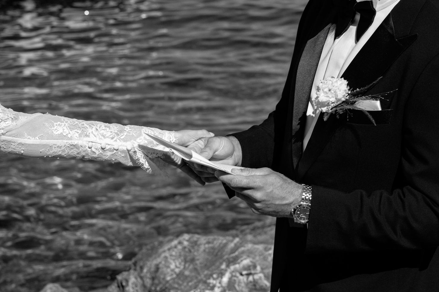 Black and white photo of hands exchanging wedding vows by the water.
