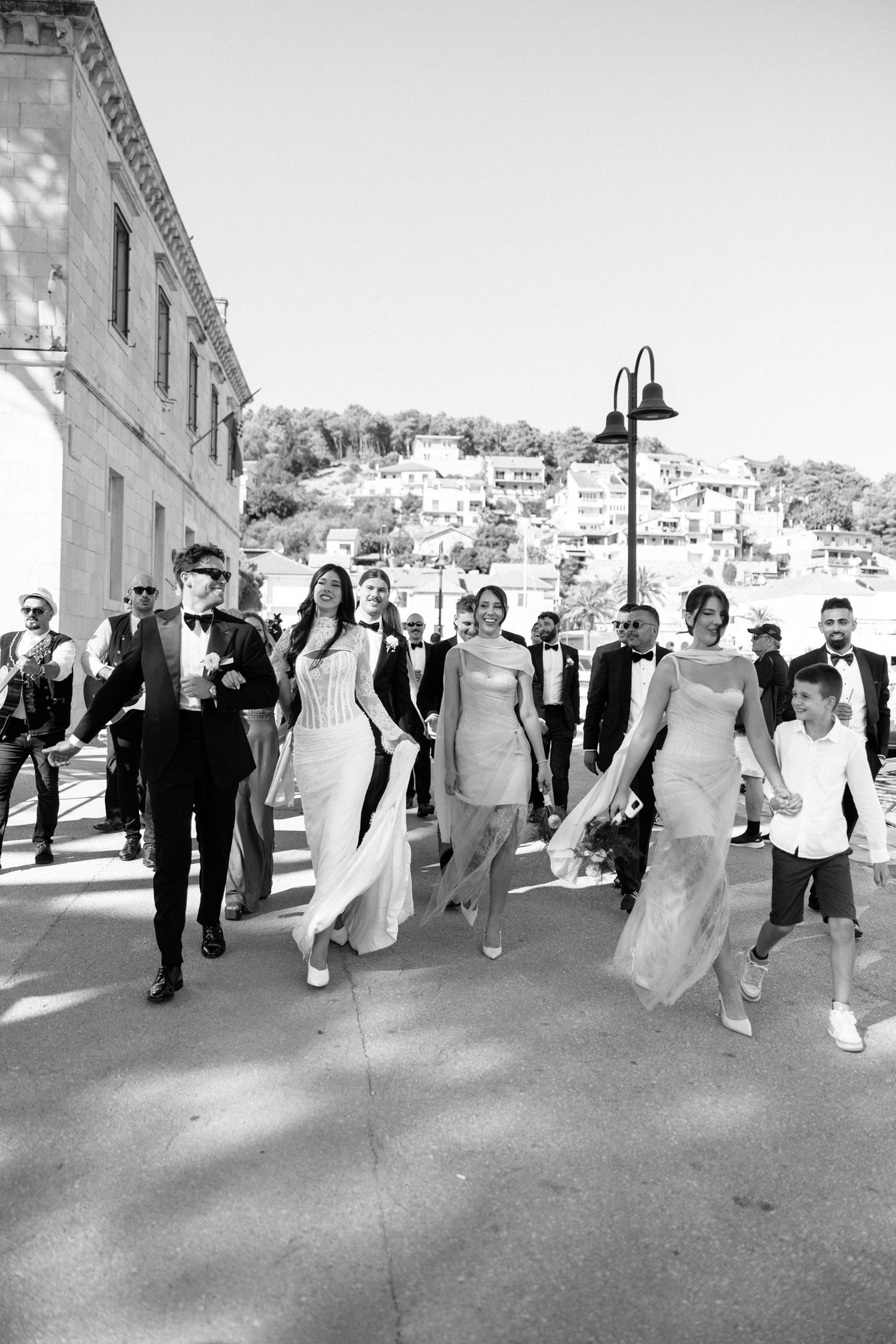 Wedding party walks down a European street with scenic hillside buildings in the background.