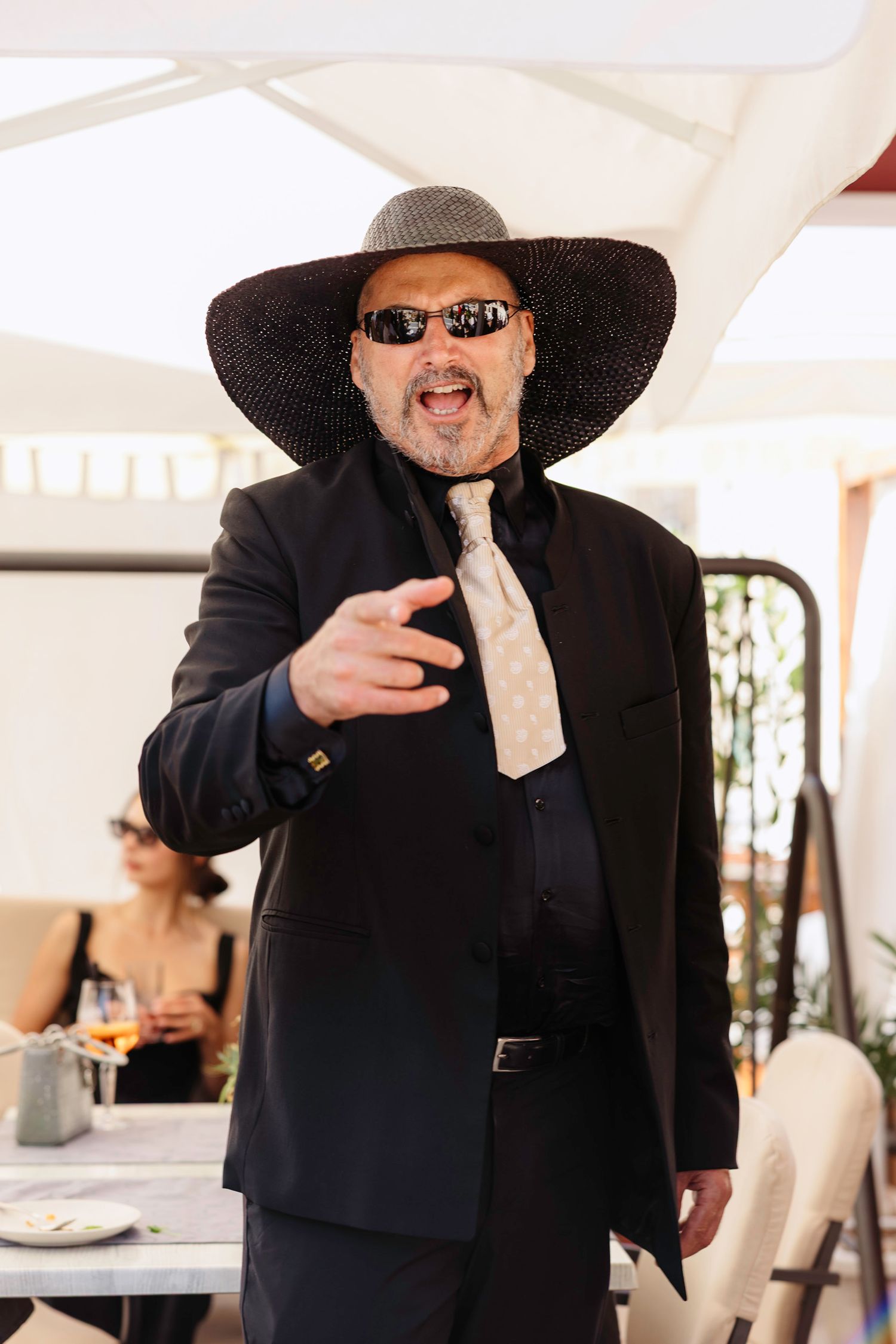 Wedding guest in a black suit and wide-brimmed hat gestures enthusiastically at an outdoor reception.