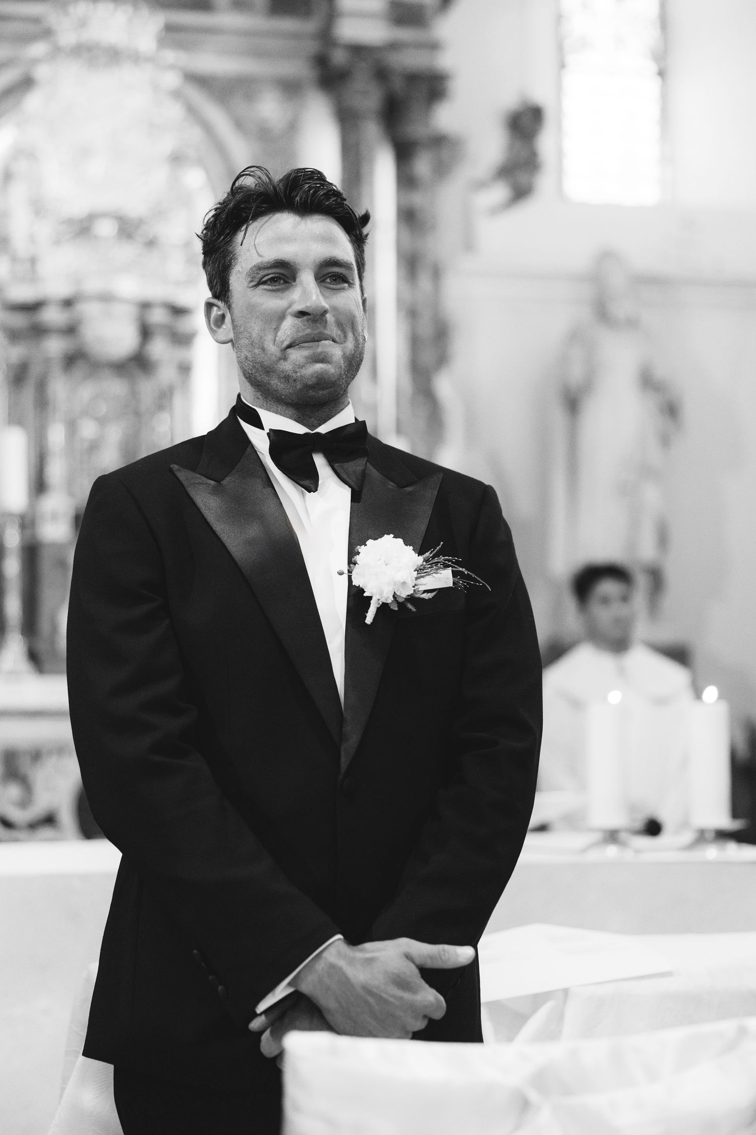A groom in a classic black tuxedo with white boutonniere stands at the altar during a church wedding ceremony.