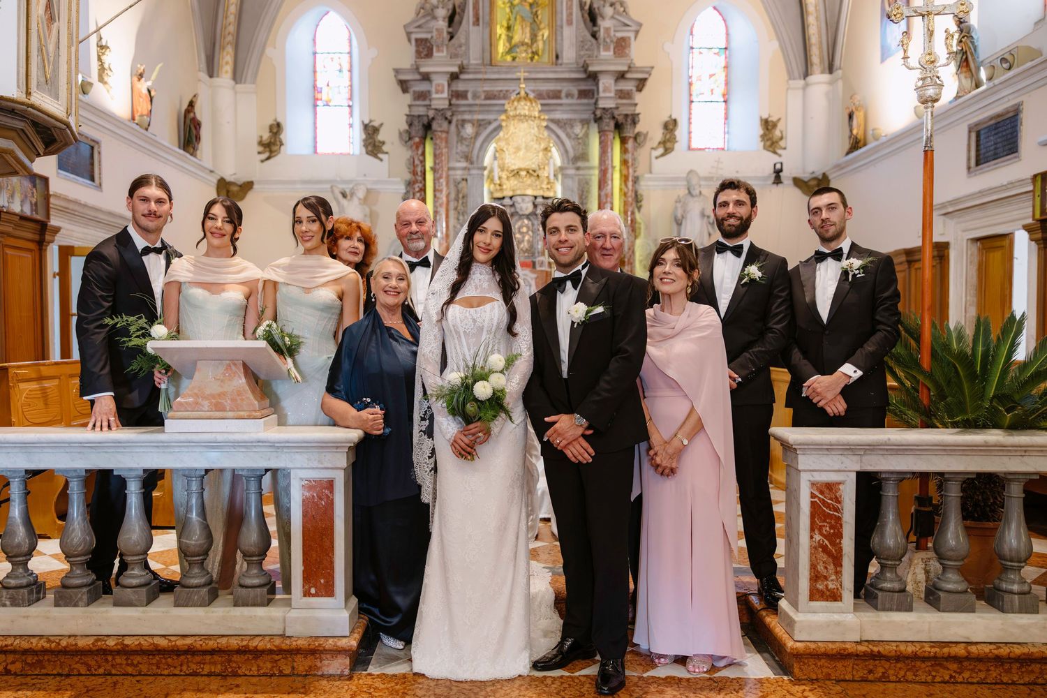 Wedding party gathered inside an ornate church with stained glass windows and elegant architectural details.