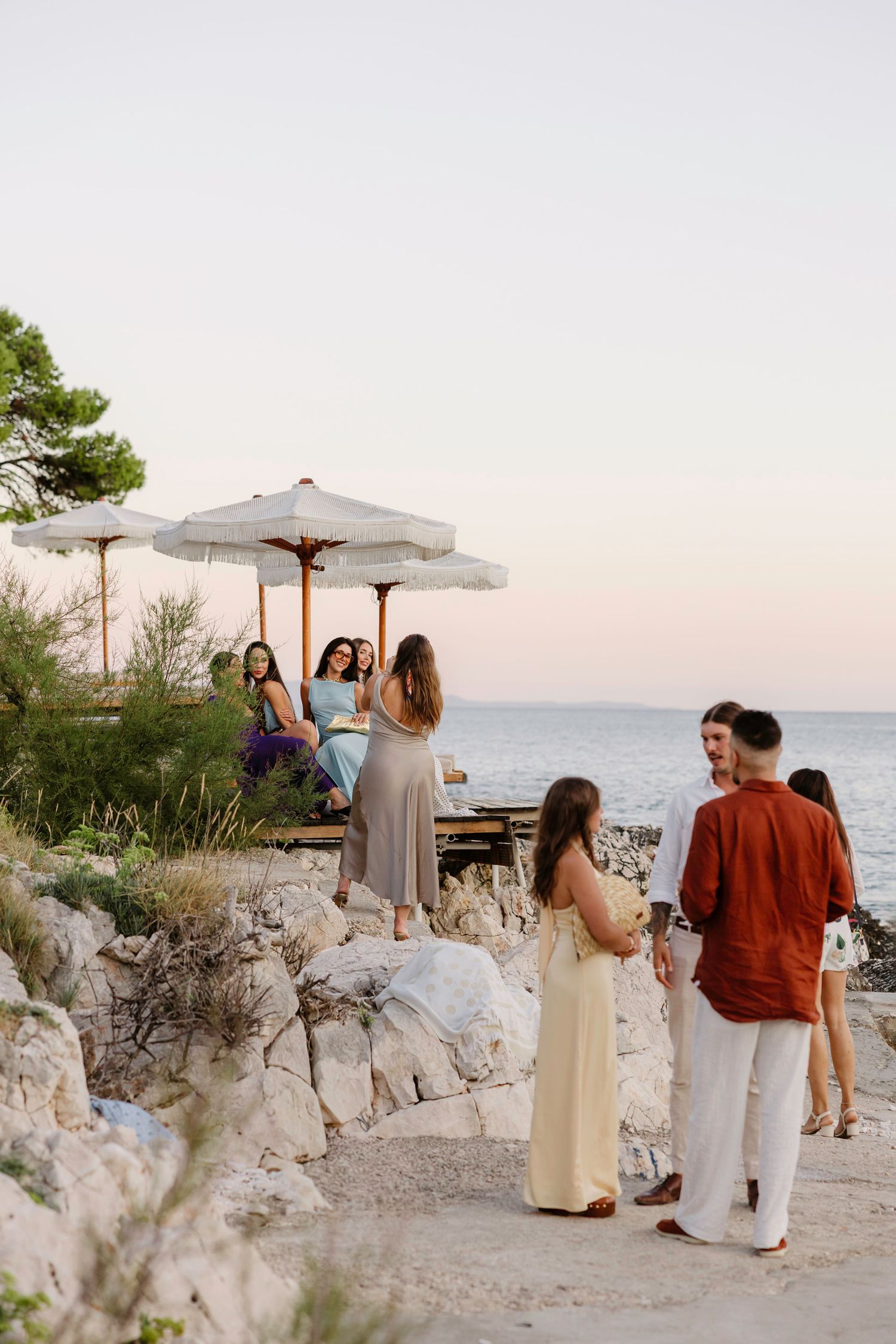 Guests gathering under white umbrellas on a rocky coastal terrace at sunset.